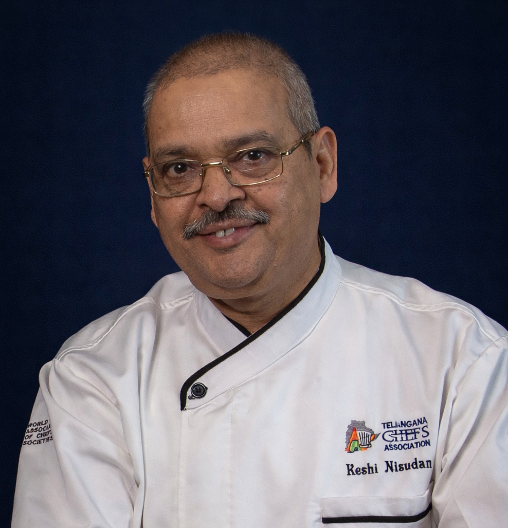 Portrait of a smiling man wearing glasses and a white chef's coat with Telangana Chief's Association logo and name tag Keshi Nisudan, against a dark background.