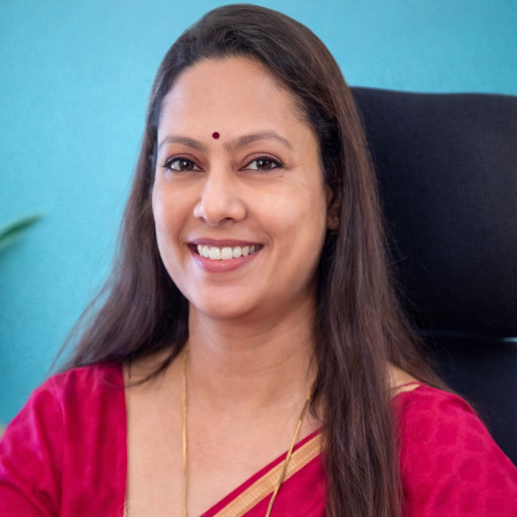 A woman smiling, wearing a red saree with gold border and a small bindi on her forehead, sitting in a black chair with a teal background.
