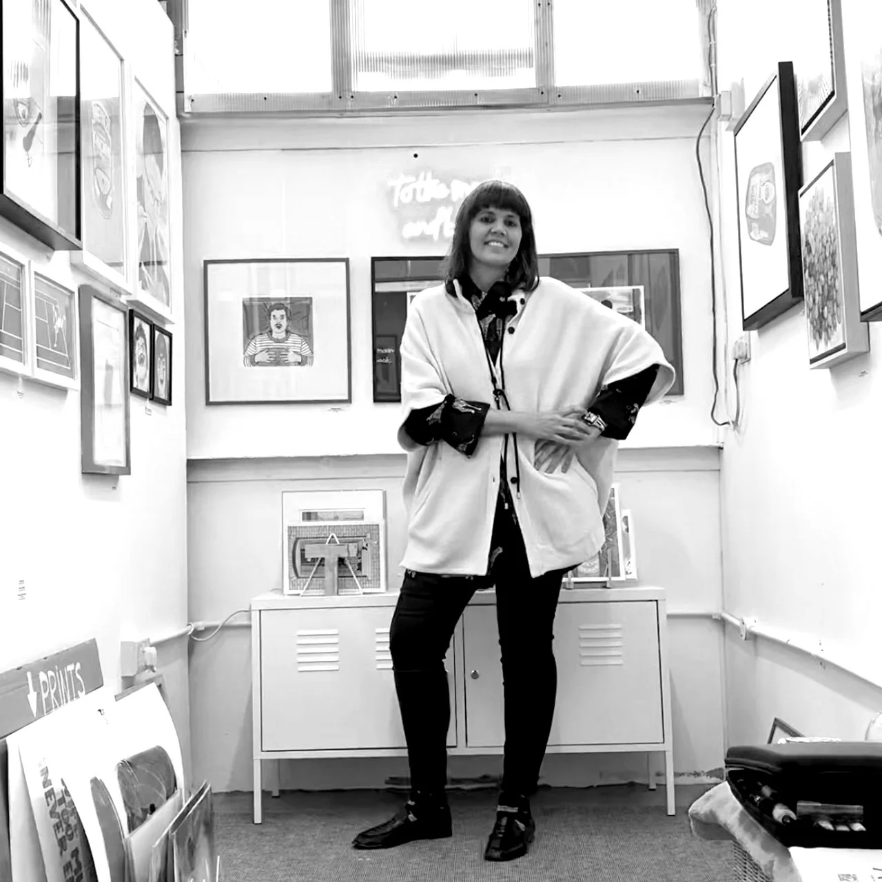 Tania Serra standing in her studio. The gallery has various framed artworks on the walls and a cabinet behind her, with prints and artworks displayed.