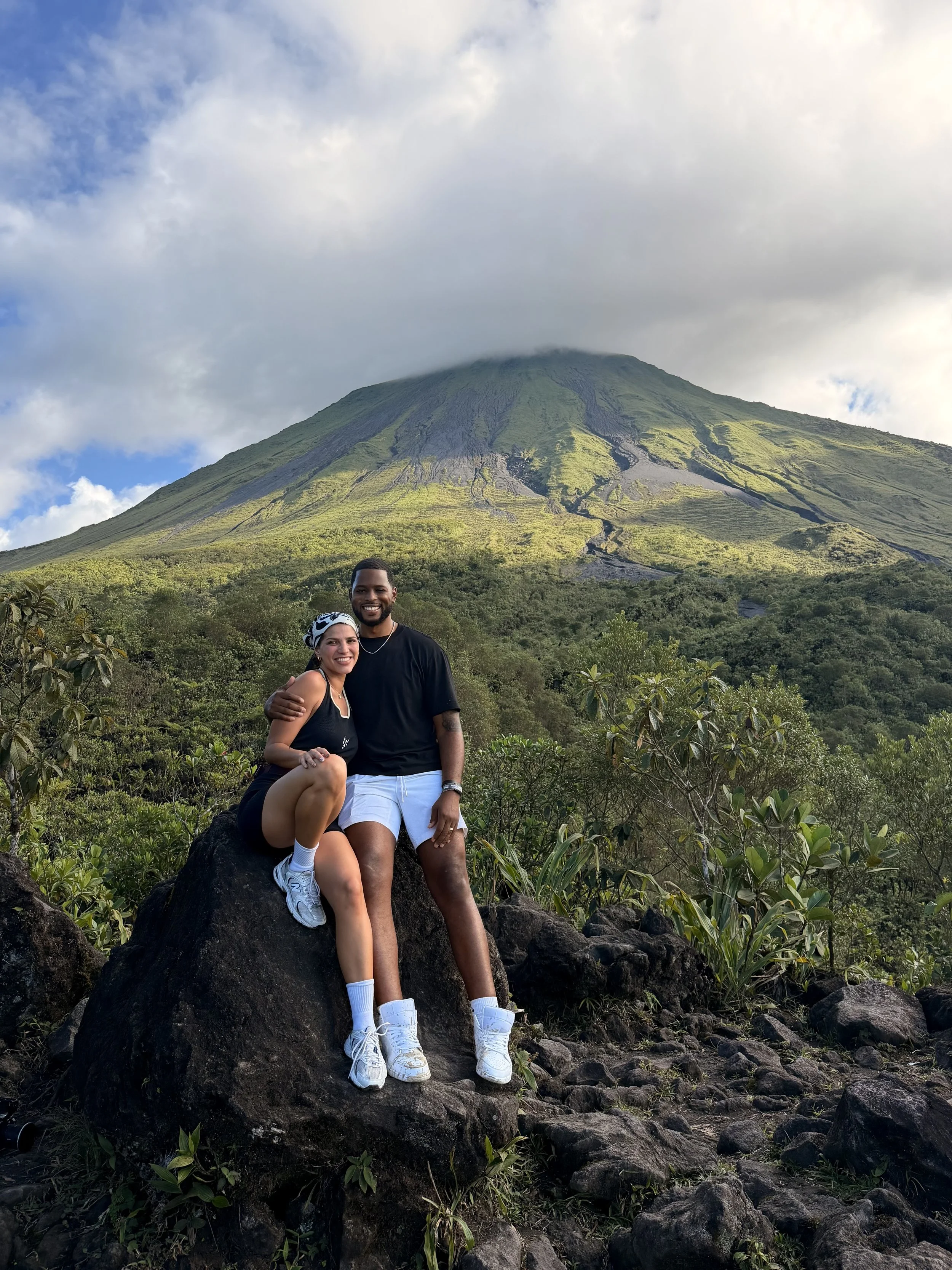A smiling couple sitting on a large rock in front of a green volcano with cloud cover at the top, surrounded by bushes and rocks.