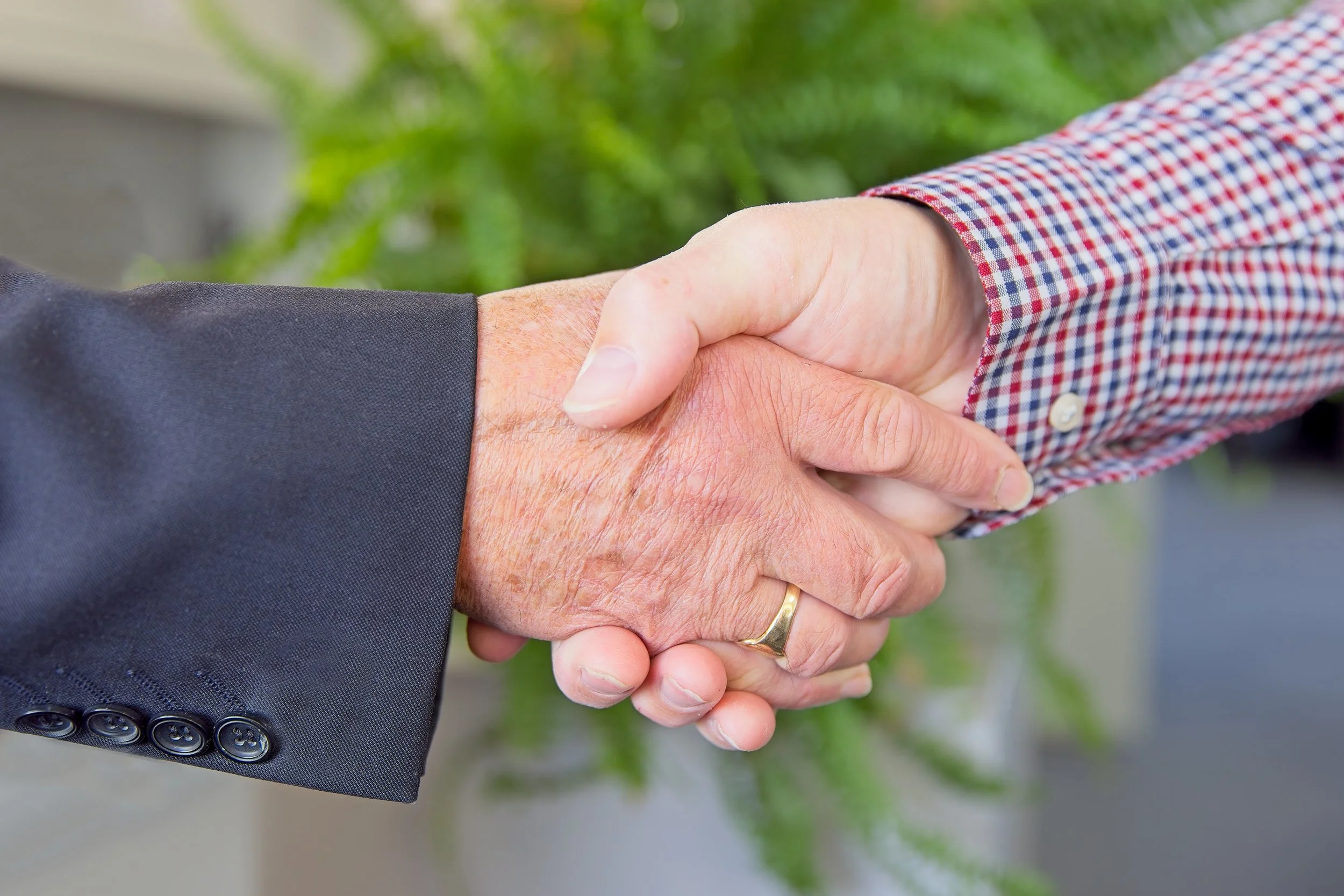 Close-up of a handshake between two men, one wearing a dark suit and the other wearing a checkered shirt with rolled-up sleeves.