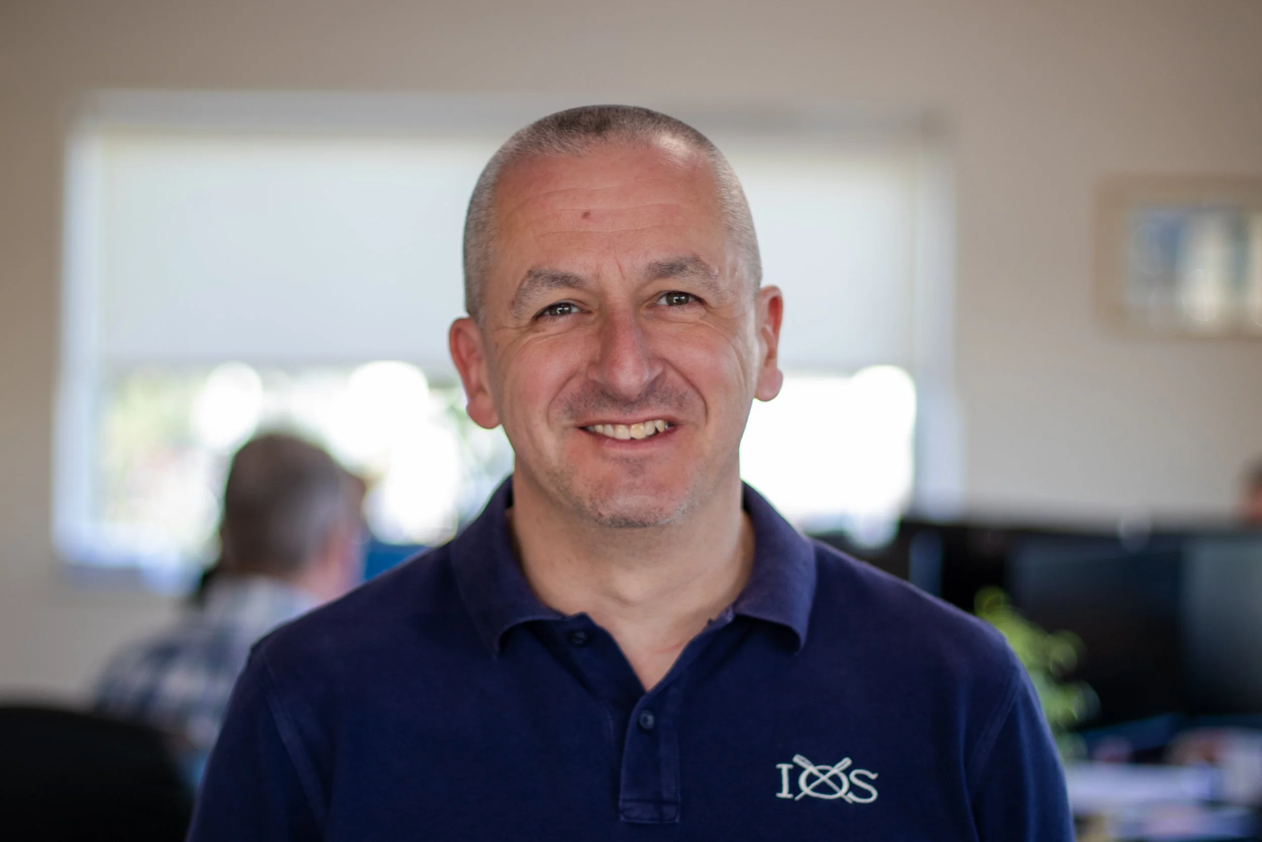 A smiling middle-aged man with short gray hair, wearing a navy blue collared shirt with a logo, standing indoors in a well-lit office setting.