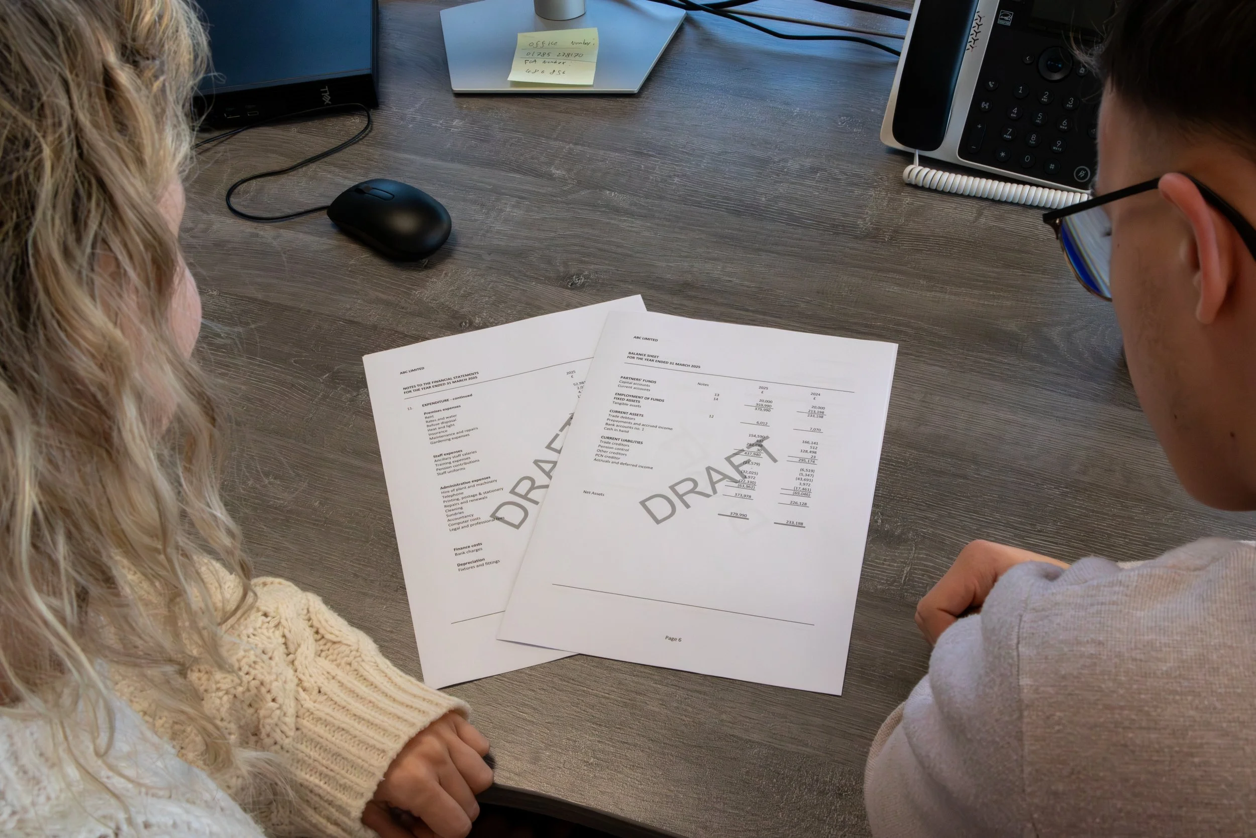 Two individuals sitting at a desk reviewing financial documents labeled 'DRAFT' with a desk phone, mouse, and sticky note on the table.