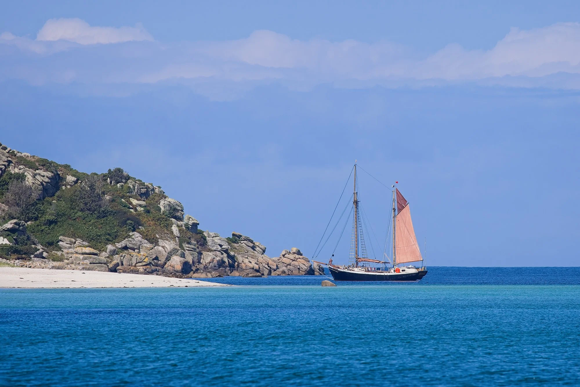 Sailboat with brown sails floating near a rocky, green-covered island on calm blue waters under a partly cloudy sky.