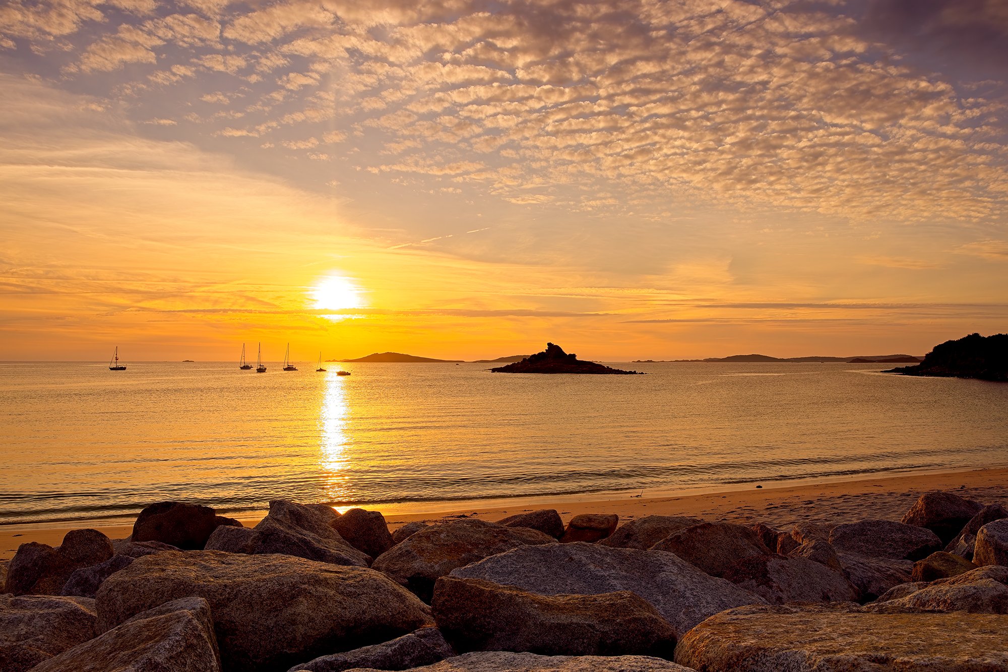 Sunset over the ocean with rocky shoreline and sailboats in the distance.