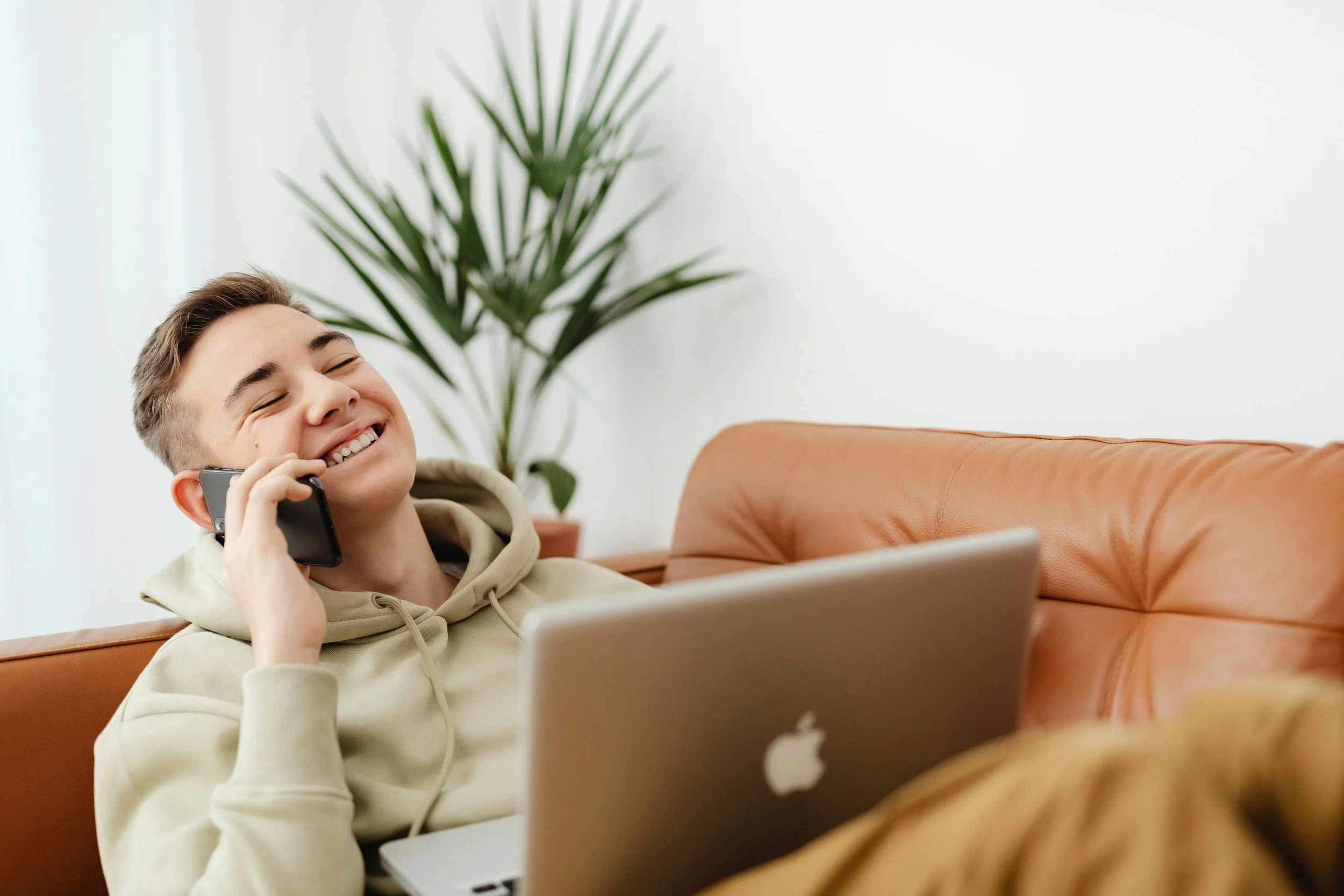 Teenage boy sitting on a couch smiling while talking on the phone with a laptop open, representing the easy next steps to starting therapy at Inside Out Therapy in Peoria Arizona