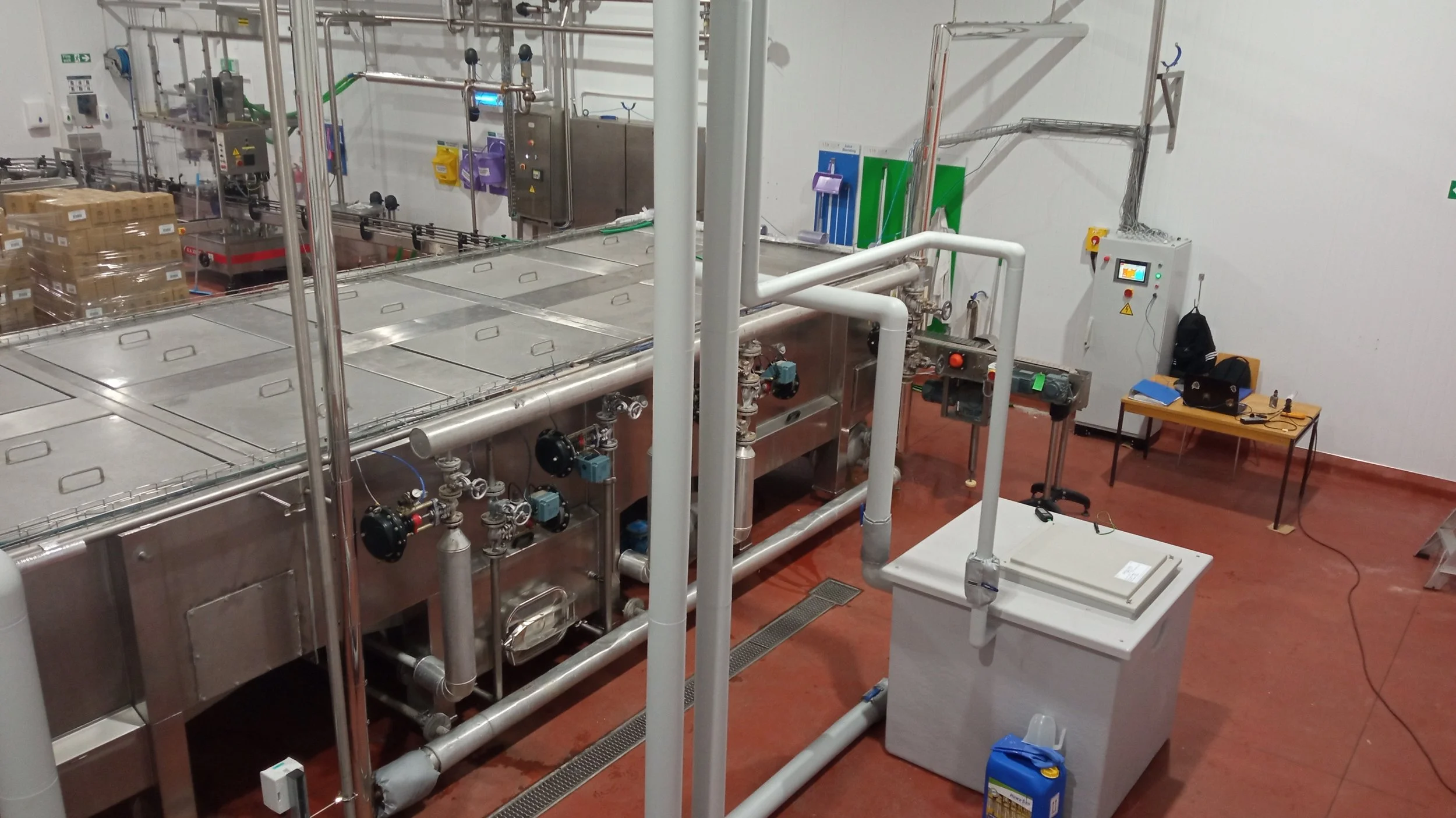 Indoor industrial laboratory or manufacturing facility with stainless steel equipment, pipes, gauges, control panels, and worktable with laptops and supplies.