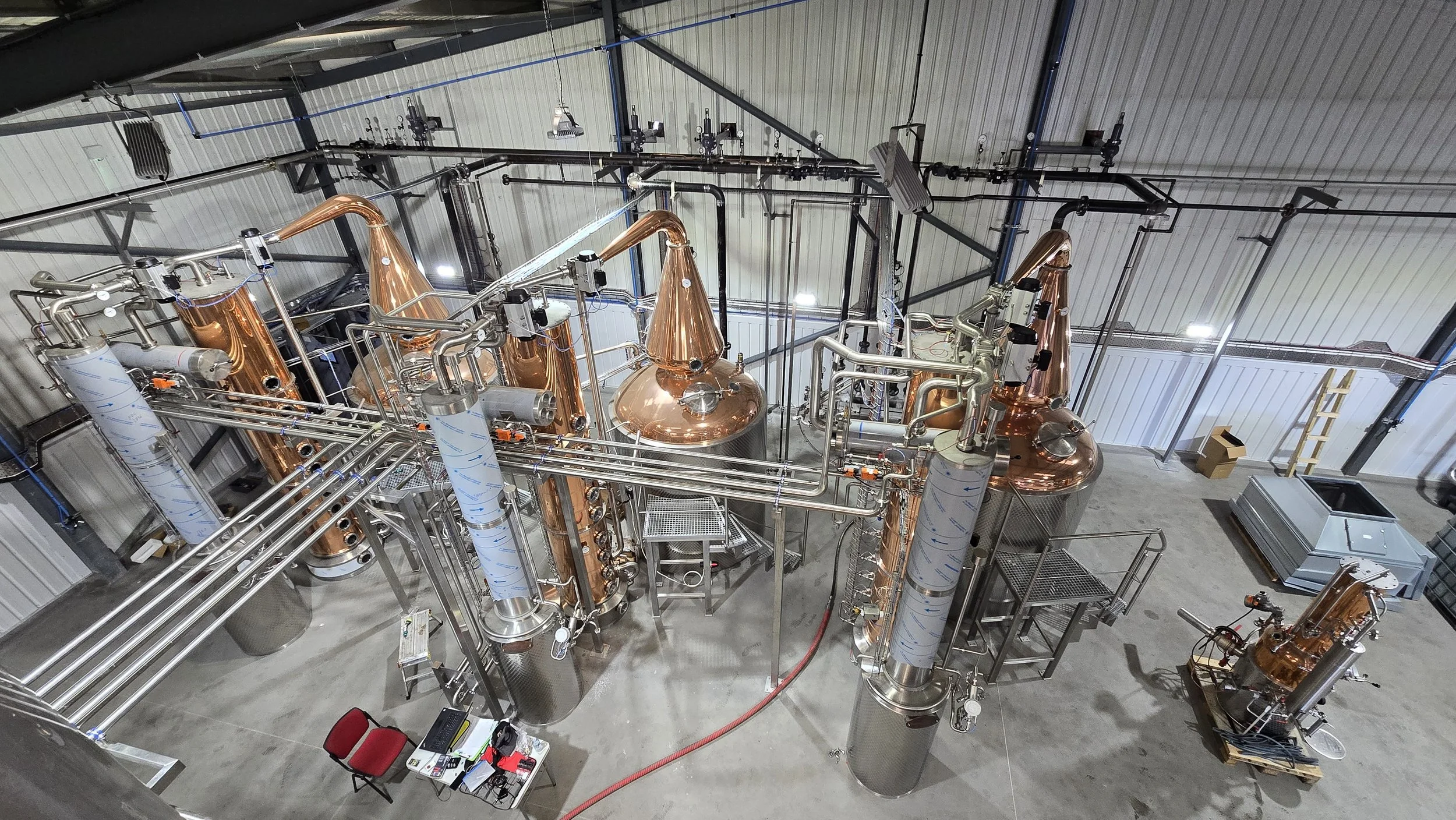 Interior view of a distillation or brewing equipment setup with stainless steel and copper vessels, pipes, control panels, and a ladder in a warehouse