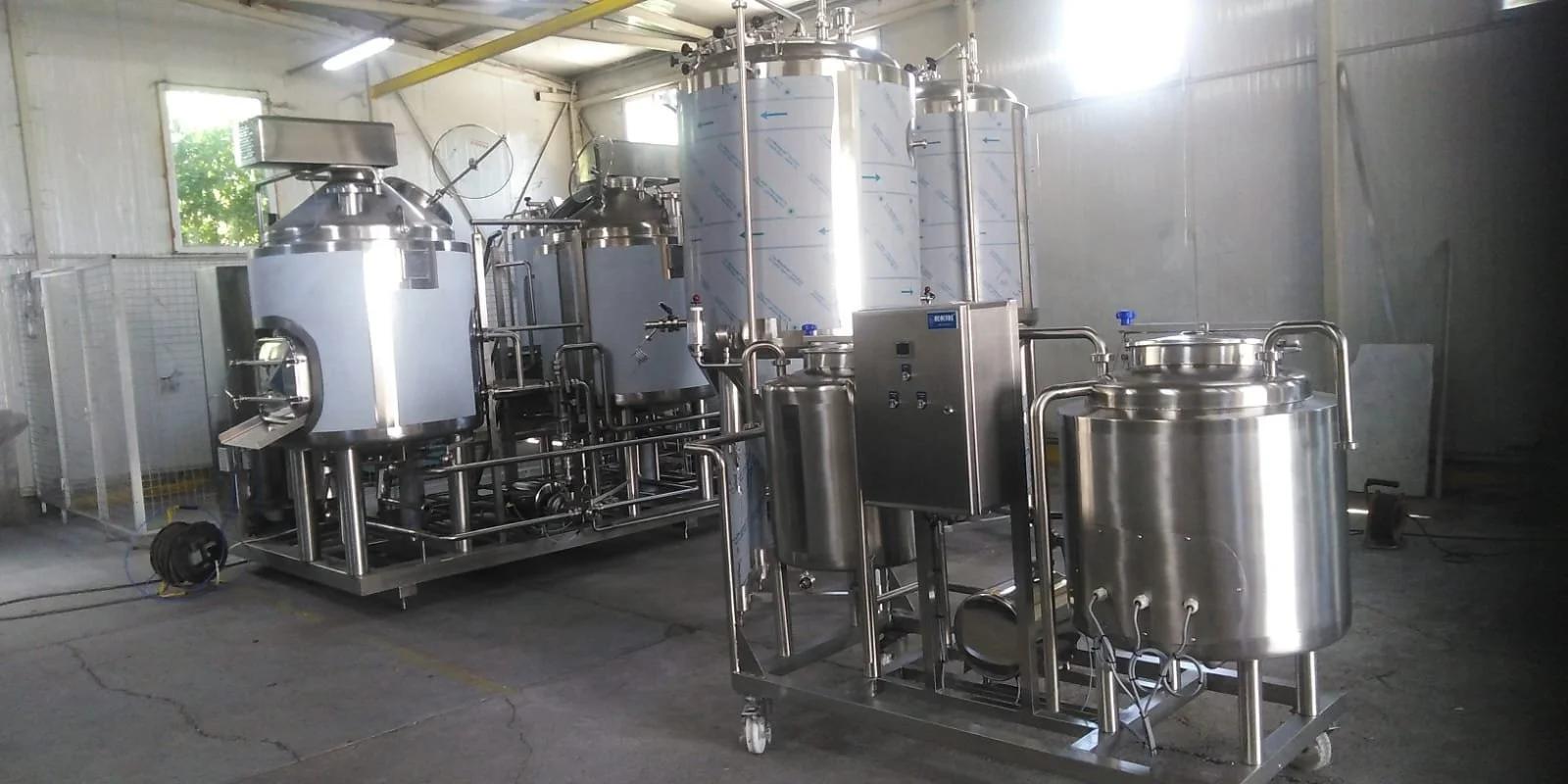 Industrial stainless steel tanks and equipment in a factory or processing plant, with pipes and control panels. Sunlight coming through window on the left.