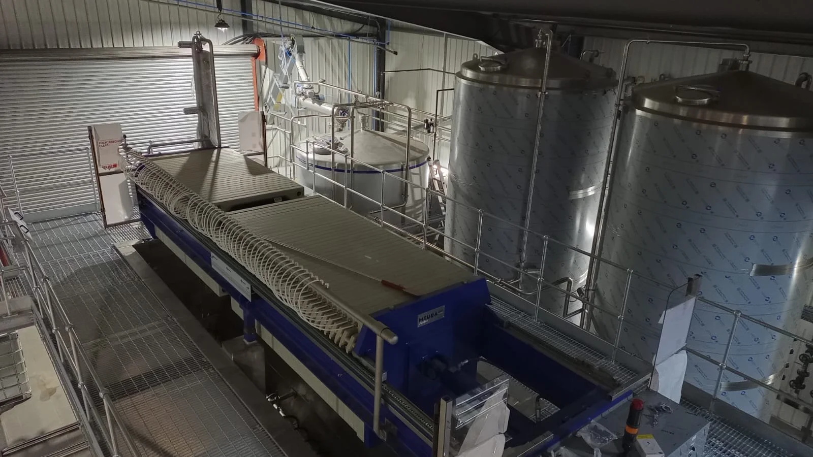 Industrial processing facility with large metal tanks and machinery, including a blue and white processing unit, surrounded by metal railings and walkways.