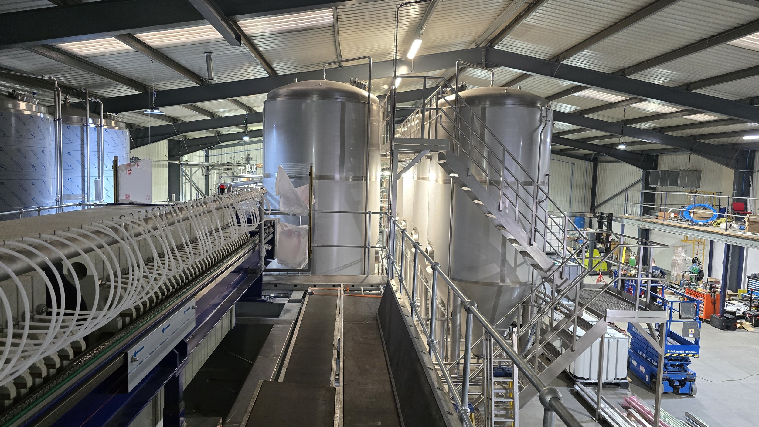 Industrial manufacturing area with large stainless steel tanks, metal stairs, conveyor system, and various tools and equipment.