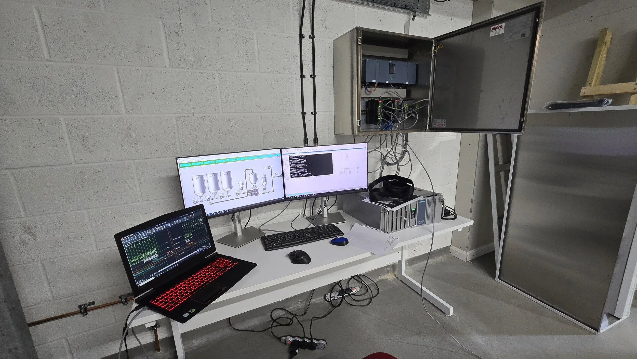 Laboratory setup with multiple computer monitors, a laptop, and electronic equipment connected to a control cabinet on the wall in a lab environment.