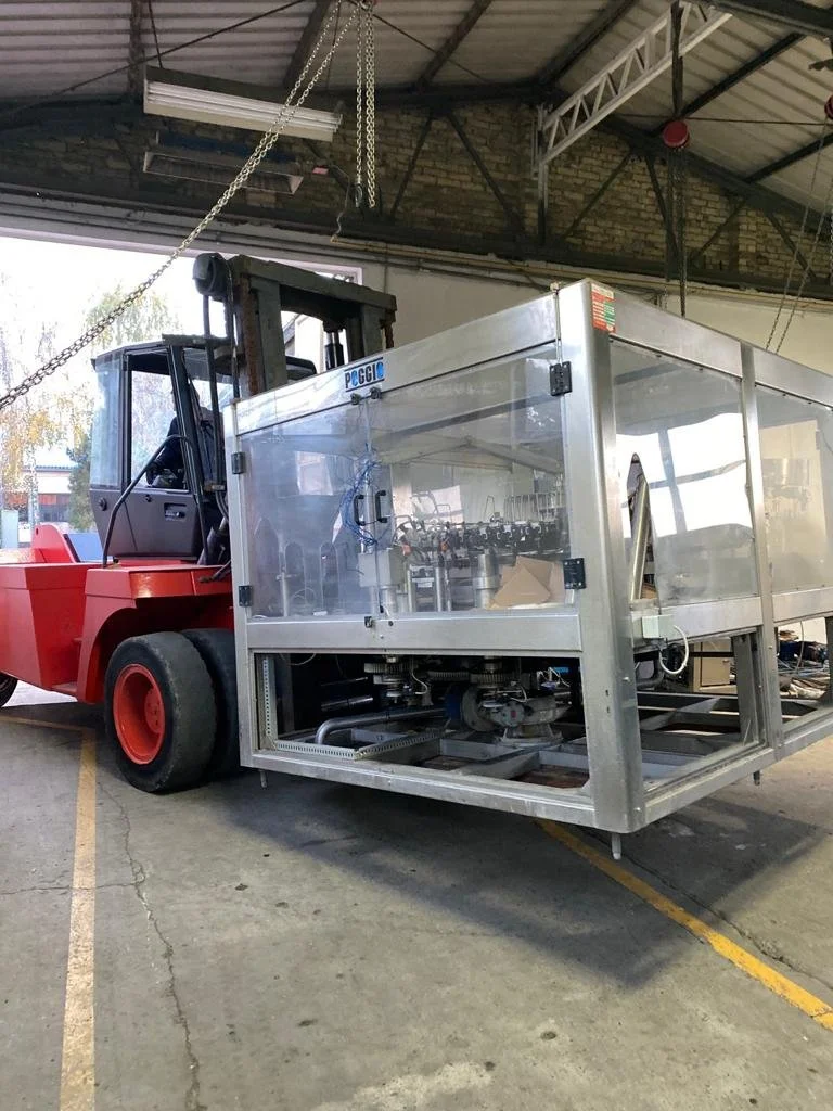 A red forklift inside a warehouse or industrial building, lifting a large metallic frame structure with transparent panels, likely part of equipment or machinery.