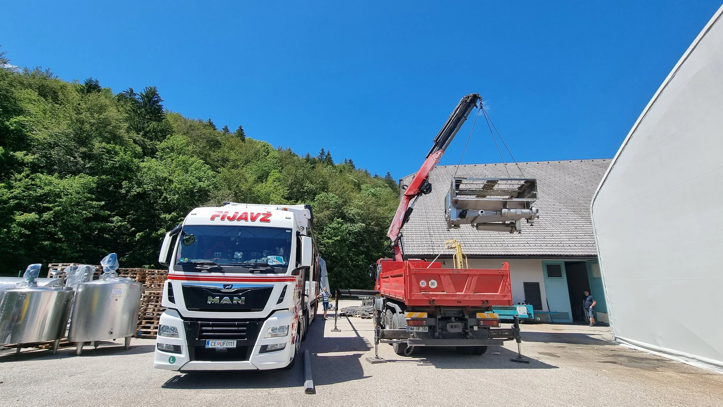 Construction site with a white truck, a crane lifting a stainless steel structure, and a building with a sloped roof, surrounded by trees and clear blue sky.