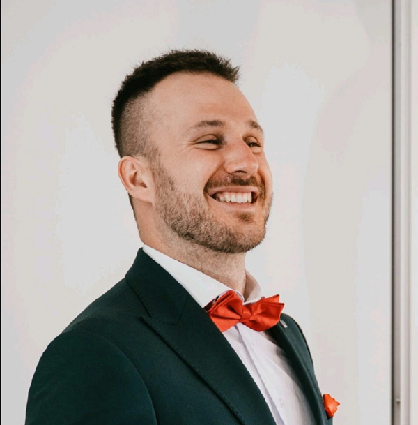 A smiling man in a suit with a red bow tie and pocket square, standing next to a metallic surface.