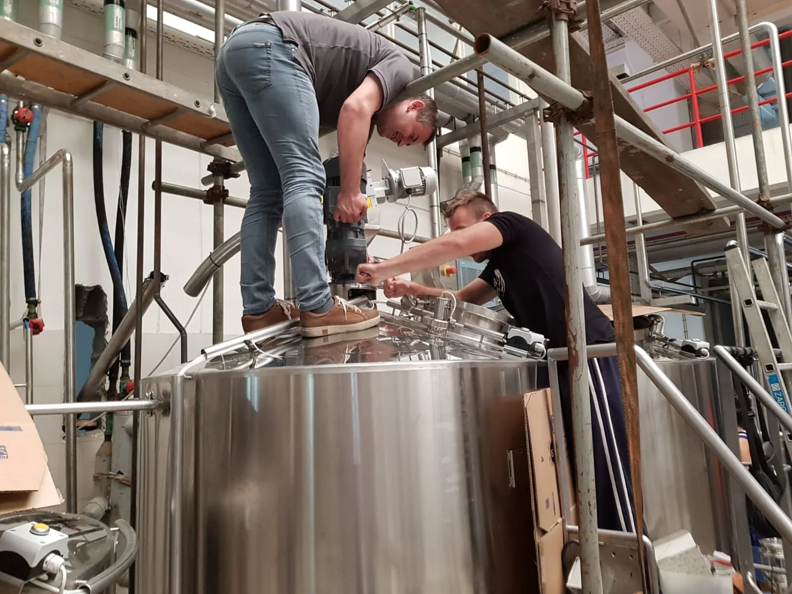 Two men working together on a large stainless steel tank, one standing on scaffolding with a power drill, the other kneeling and holding parts of the tank.