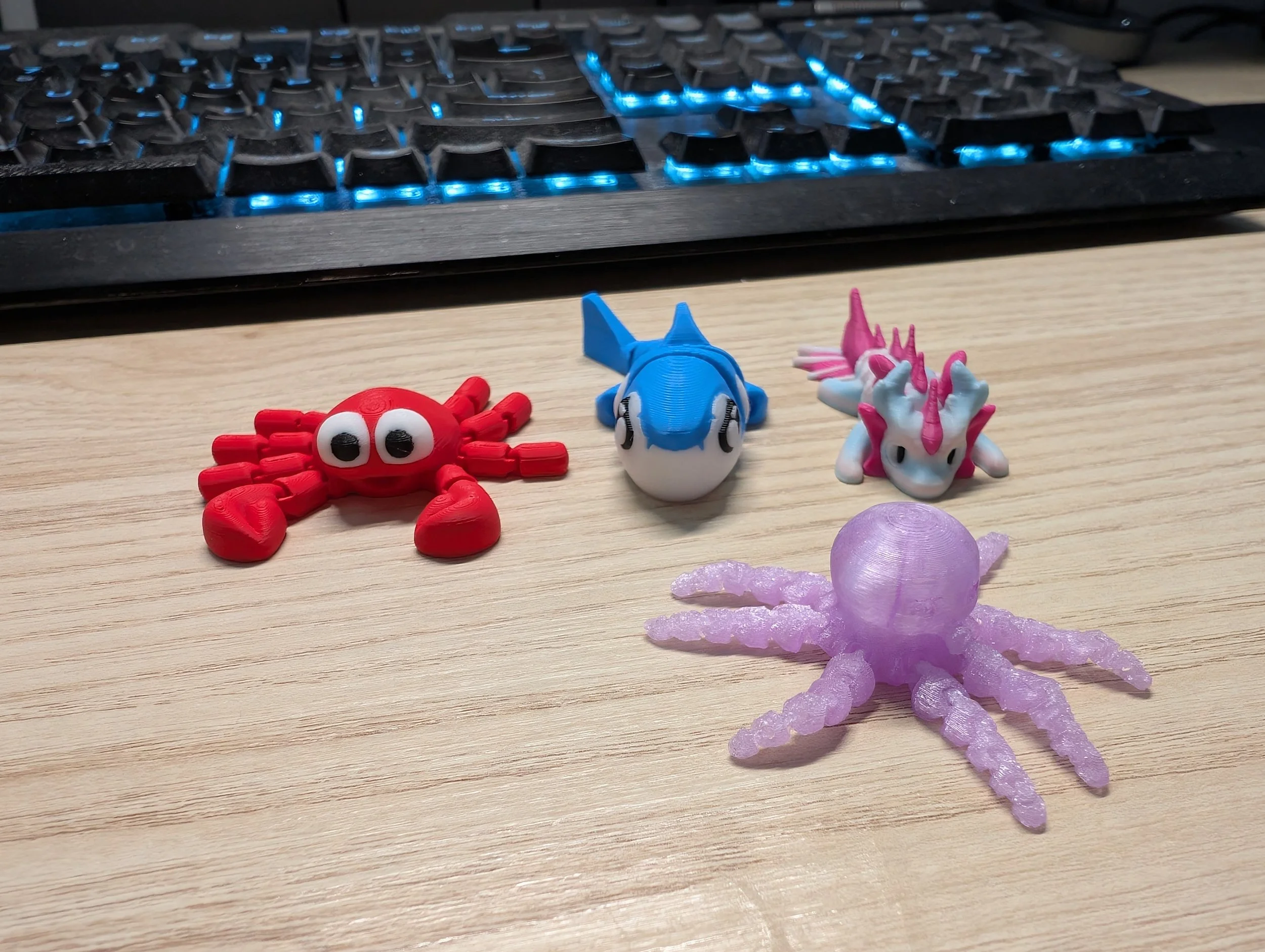 Collection of four small toy creatures including a red crab, a blue fish, a pink and white unicorn, and a purple octopus on a wooden surface with a keyboard in the background.