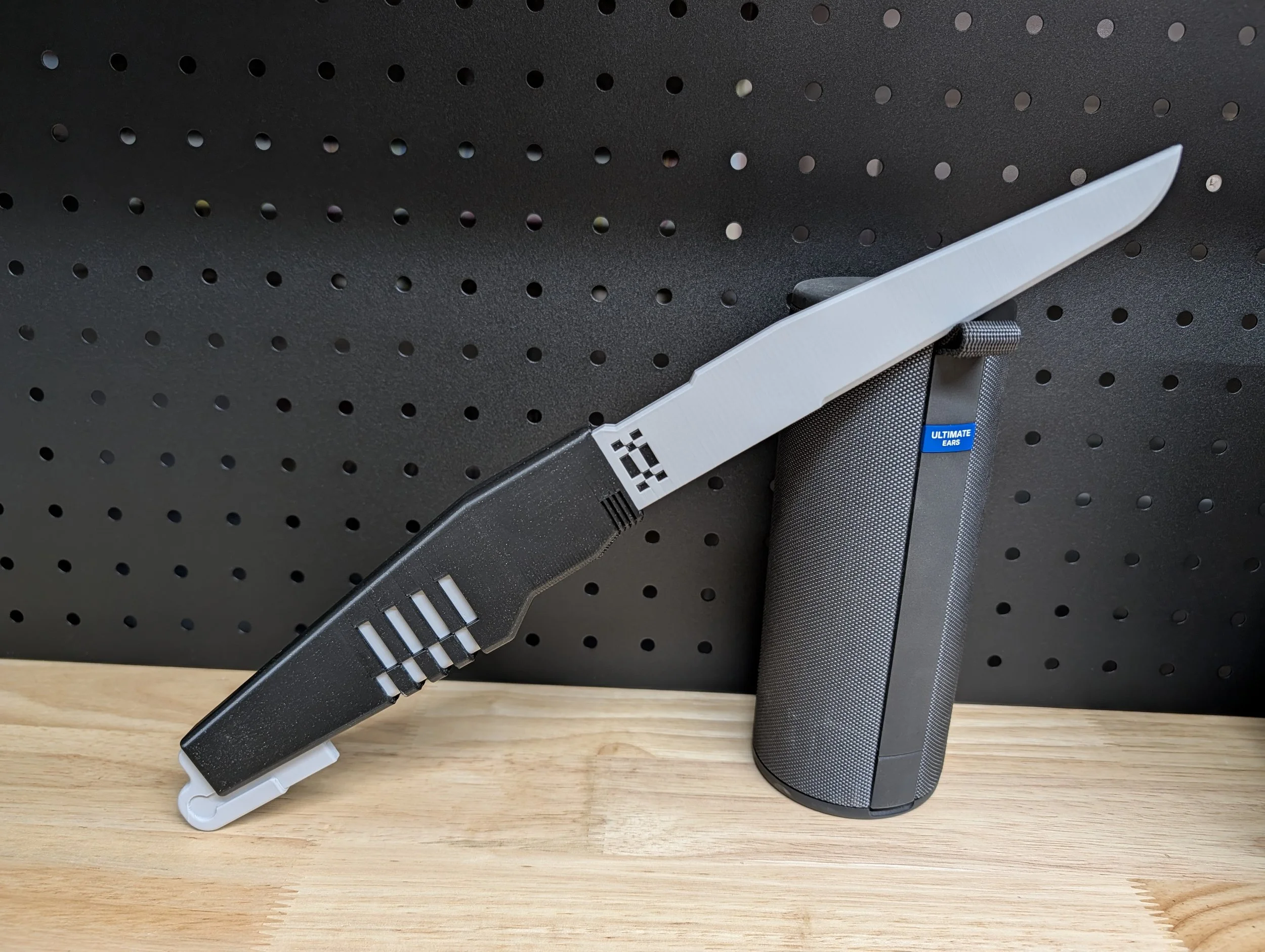 A large black and white folding knife displayed against a pegboard wall, with a cylindrical gray speaker positioned behind it on a wooden surface.