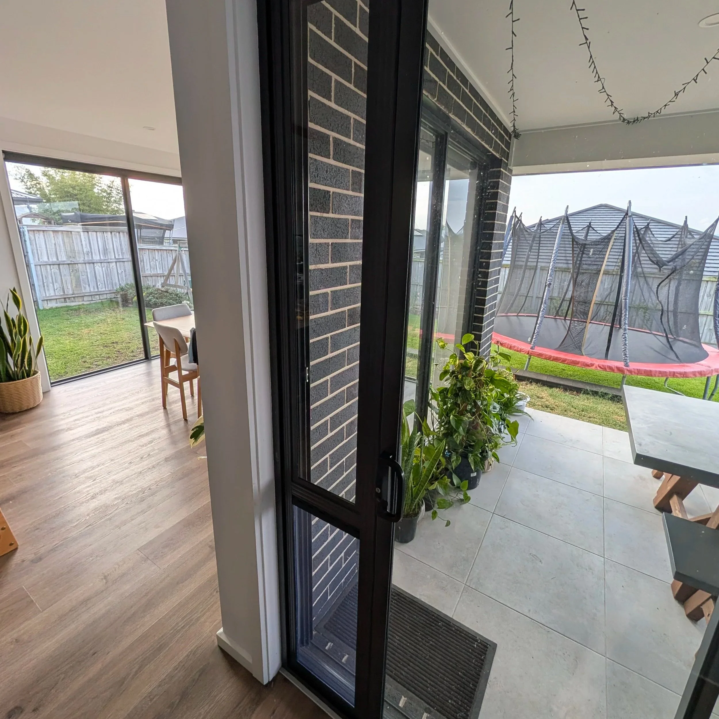 View of a screened porch with plants, a table, and a trampoline outside. The porch has tiled flooring and black framed sliding glass doors.