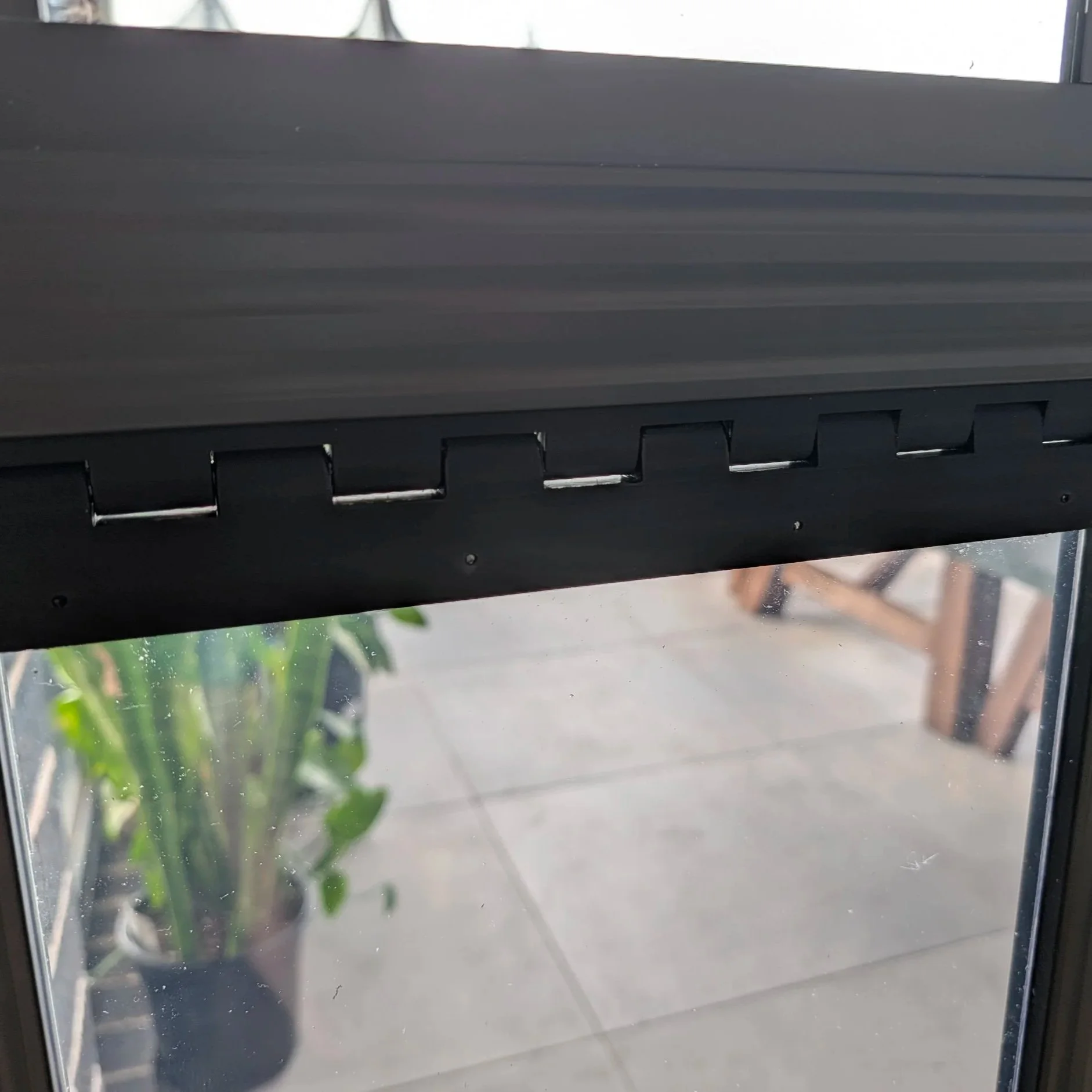 Close-up of a black roller shade partially lowered on a window, with a potted plant and patio visible outside.