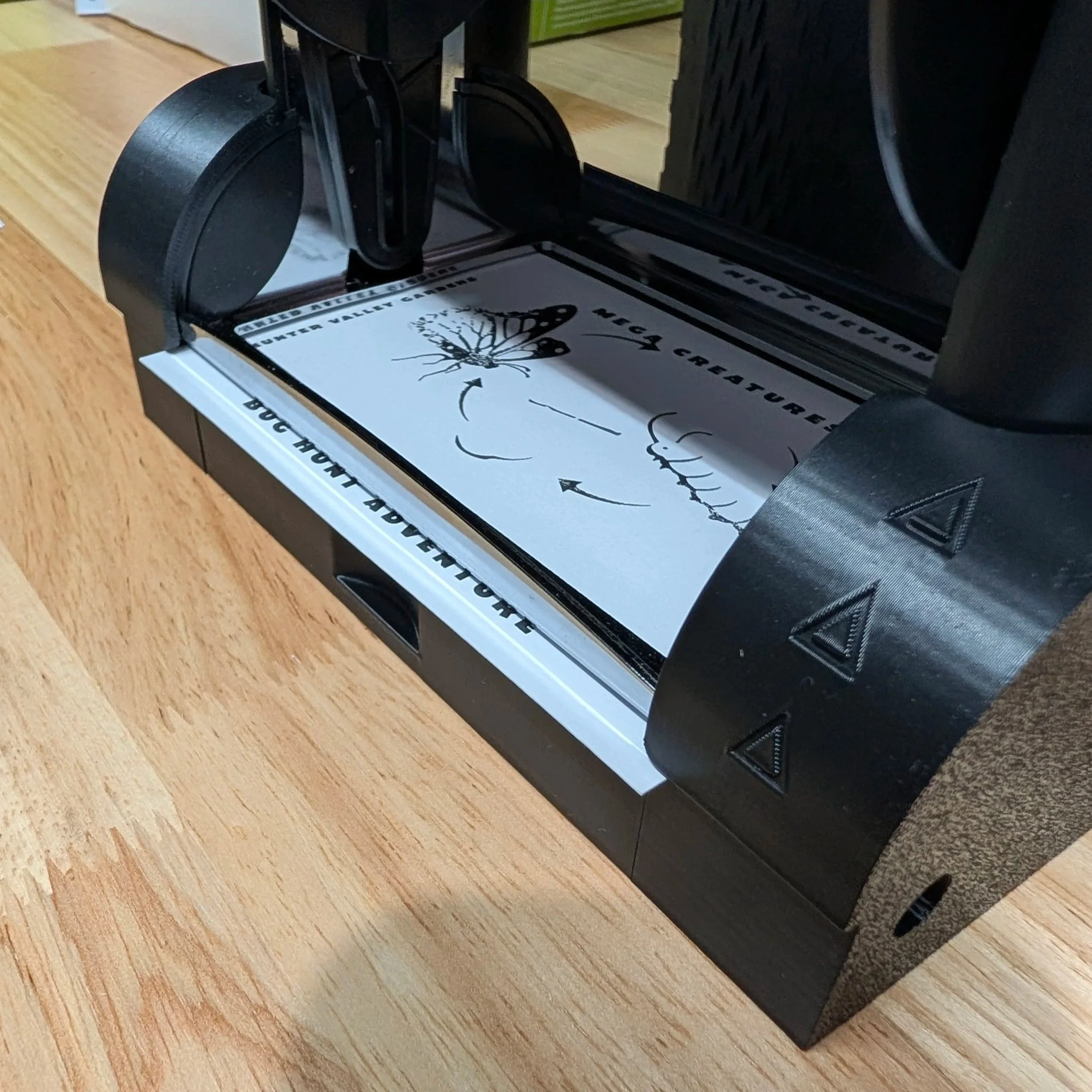 A small black laser cutter or engraver machine on a wooden surface, with paper inside featuring a diagram of a butterfly and arrows, designed for creating or etching detailed designs.