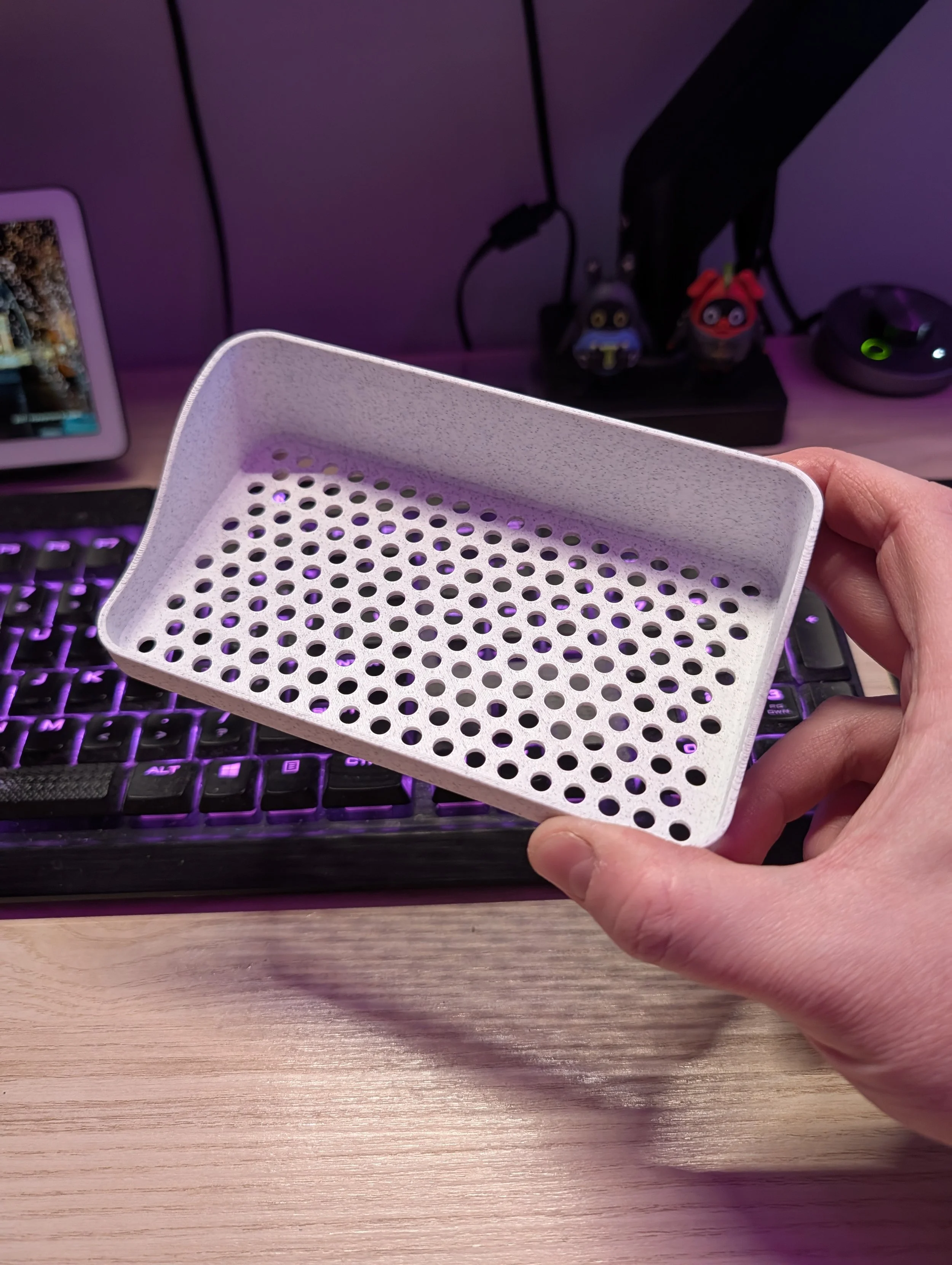 Person holding a small white perforated plastic tray in front of a computer keyboard with purple backlight on a wooden desk.
