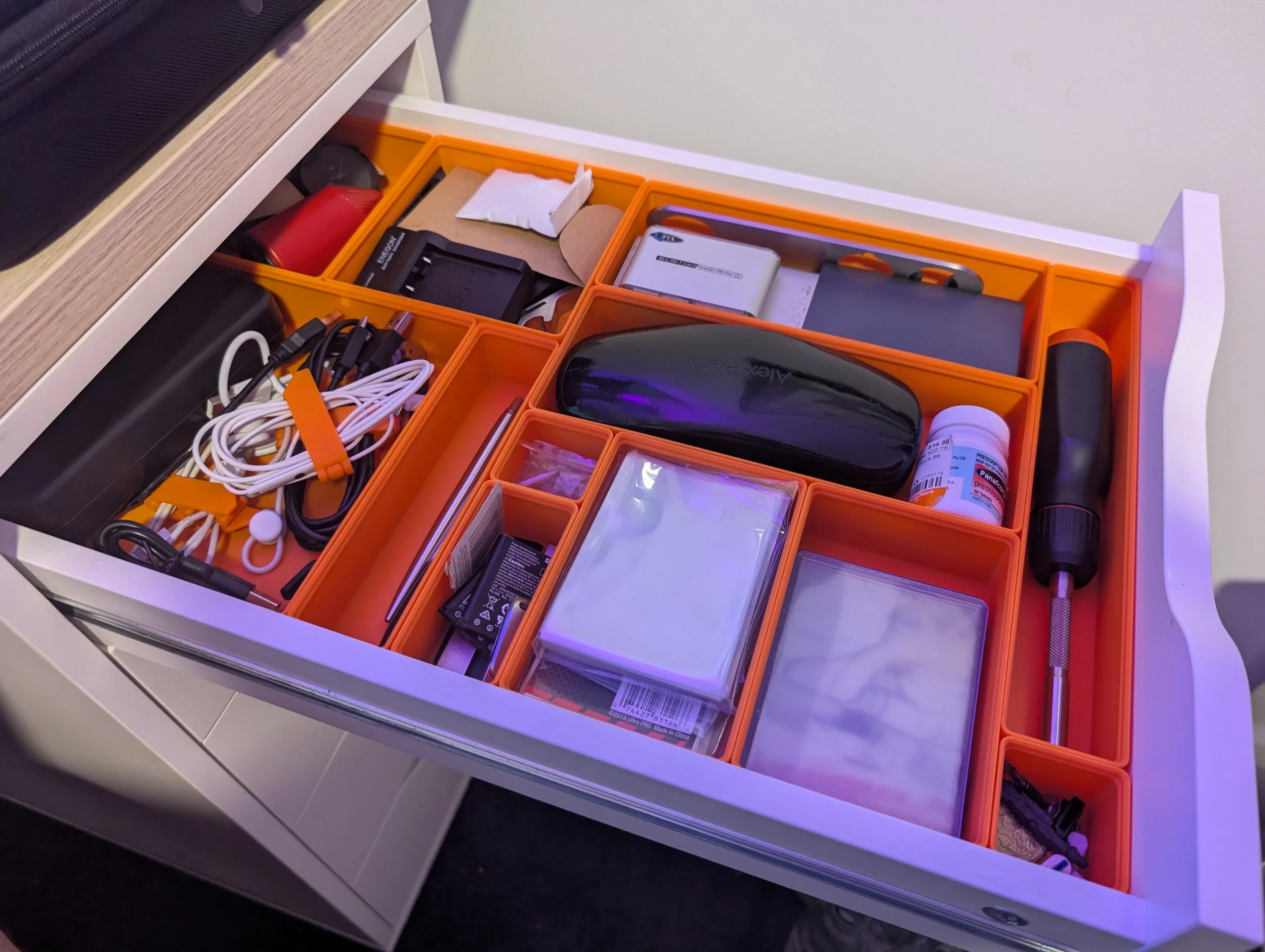 Open drawer with organized orange compartments containing electronic accessories, cables, a black case, medication pills, and small tools.