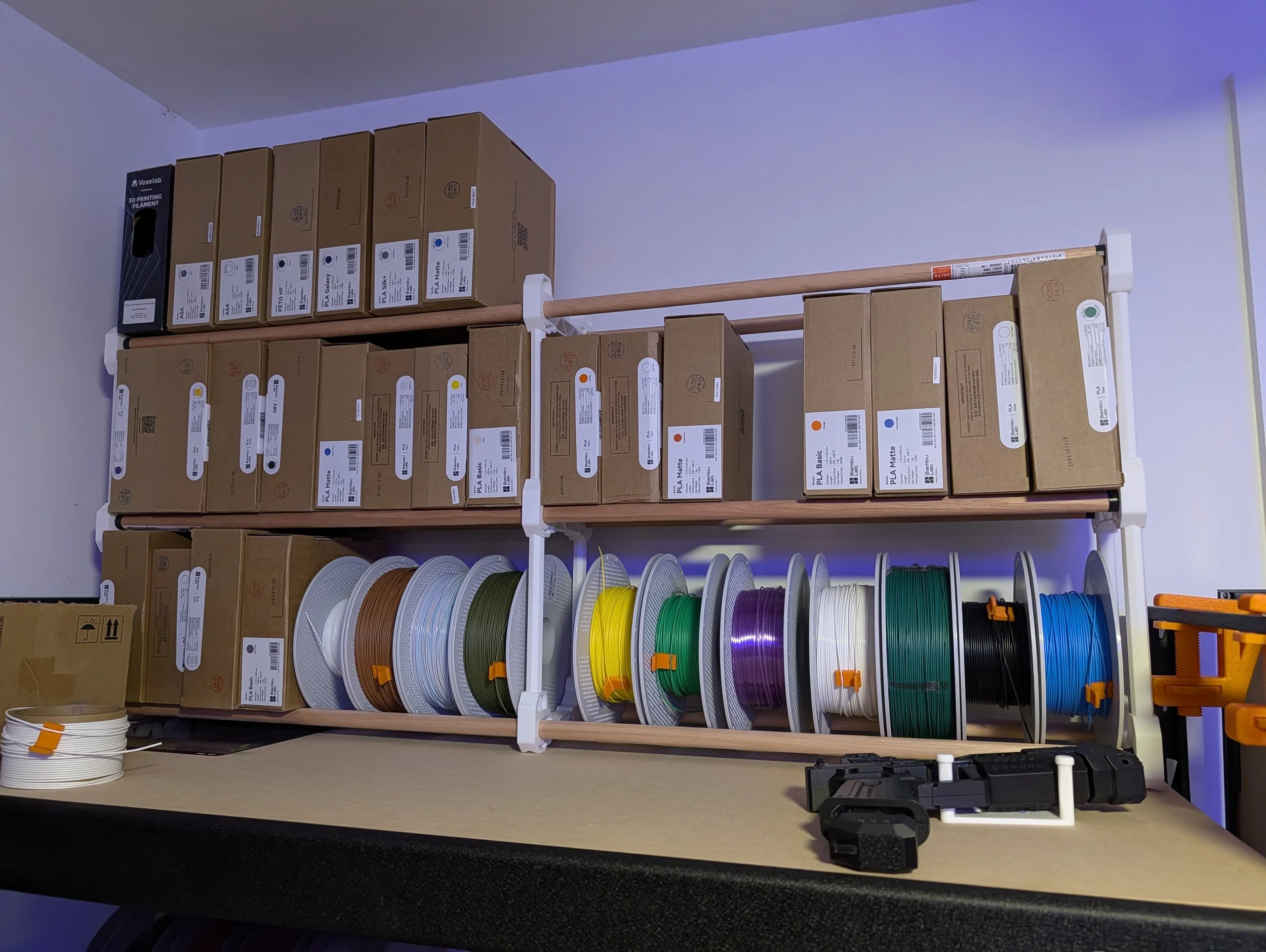 Shelves with boxes of filament spools in various colors, including white, black, green, purple, yellow, and blue, used for 3D printing, with tools and a cardboard box on the table below.