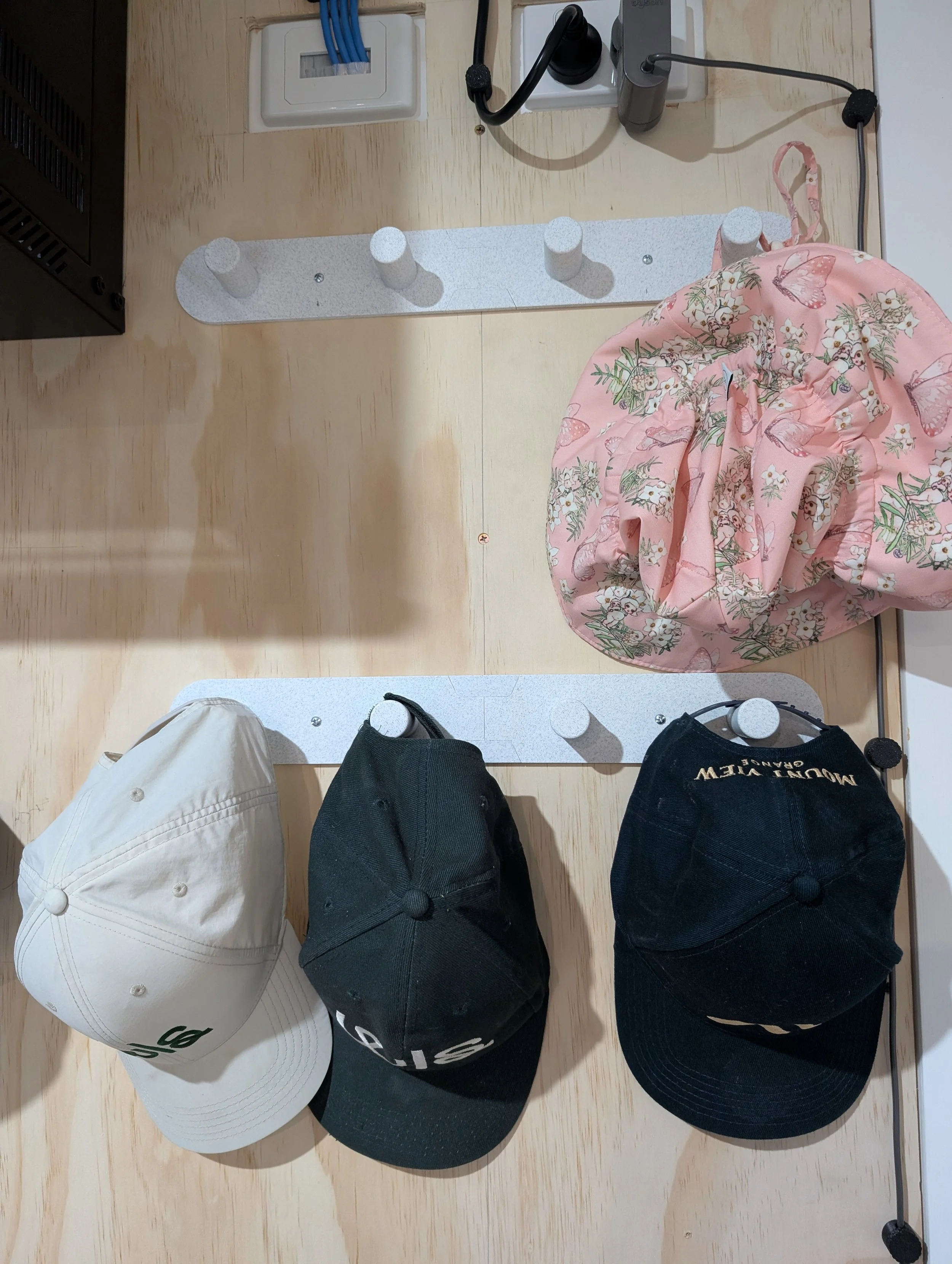 Three baseball caps hanging on a wall hooks, with a pink floral bag on the right.