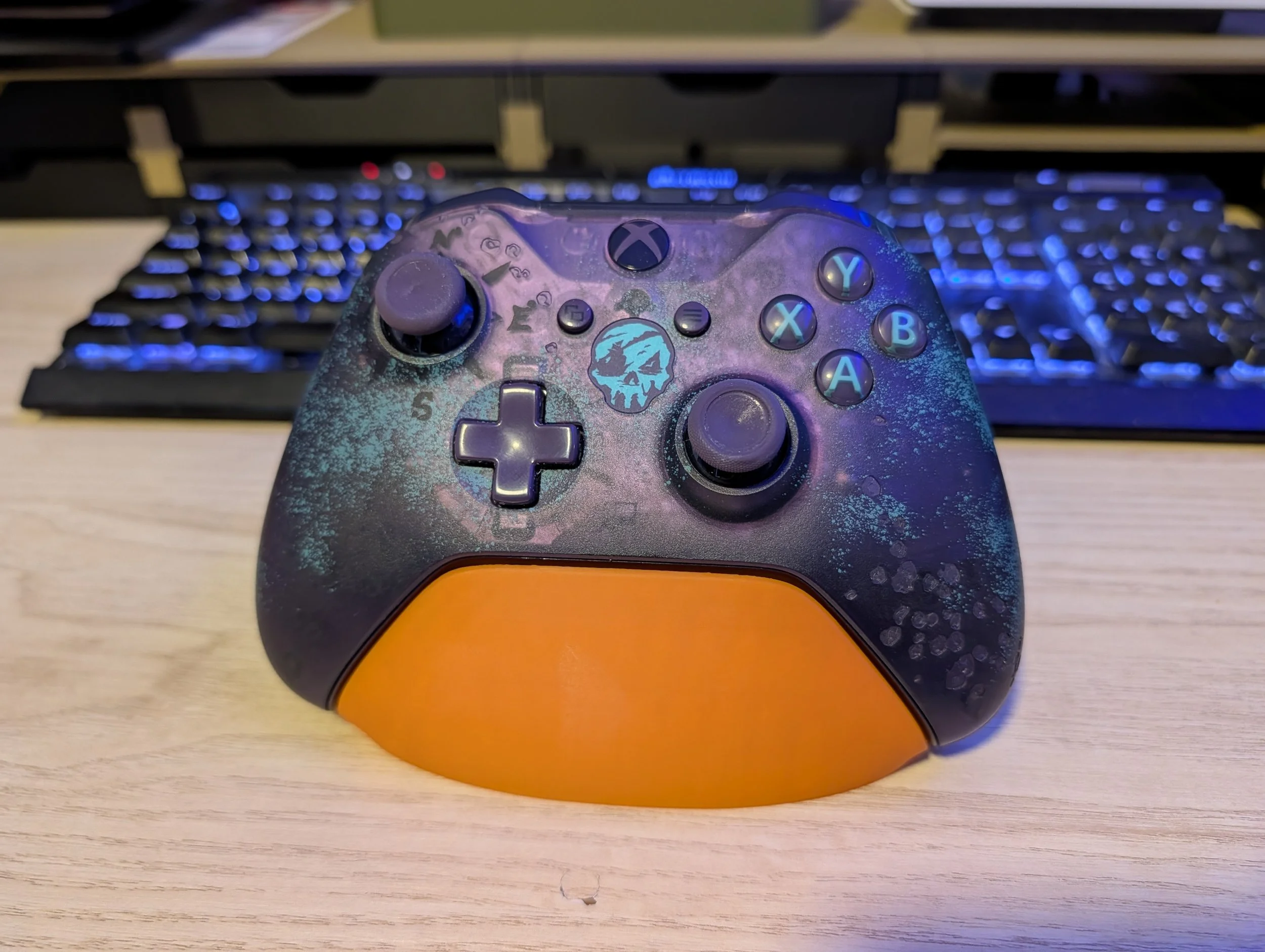 A gaming controller with a splattered purple and blue design and an orange bottom section, placed on a wooden desk with a blurred keyboard in the background.