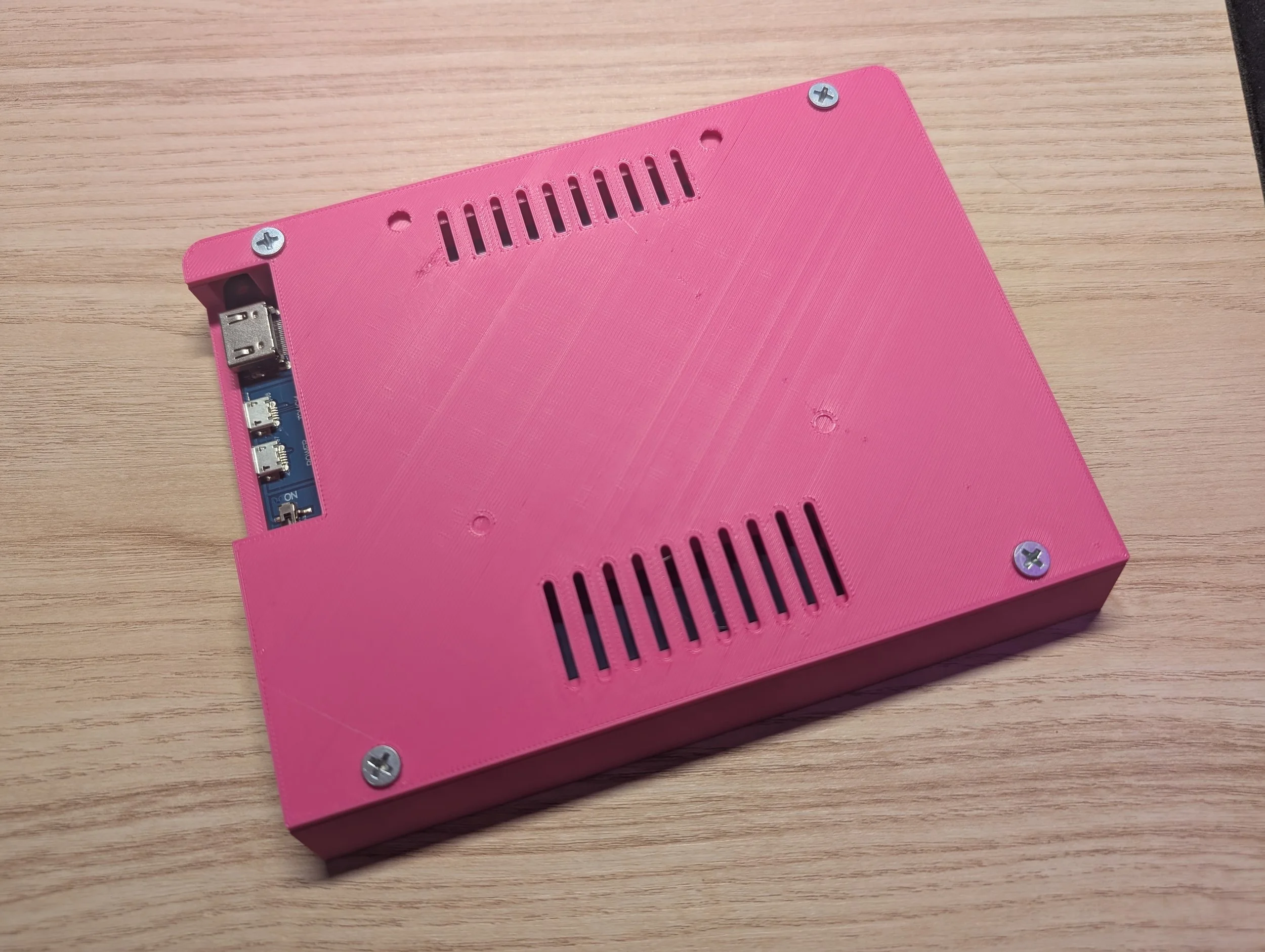 Pink electronic enclosure with vent holes and visible USB and GPIO ports on a wooden surface.