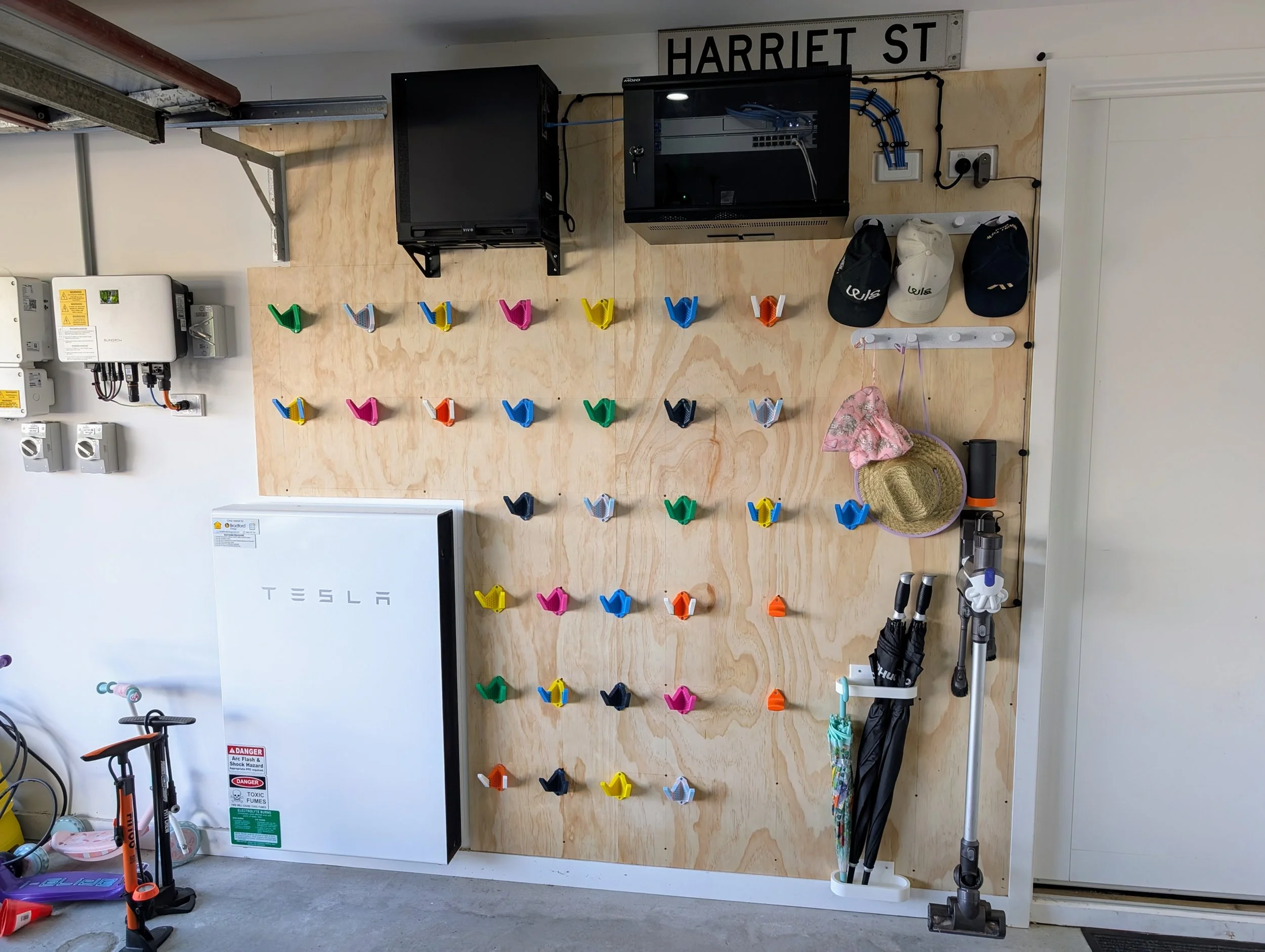 Garage wall with a pegboard featuring colorful climbing holds, a Tesla refrigerator, various hats, a baseball helmet, an umbrella, and a vacuum cleaner.