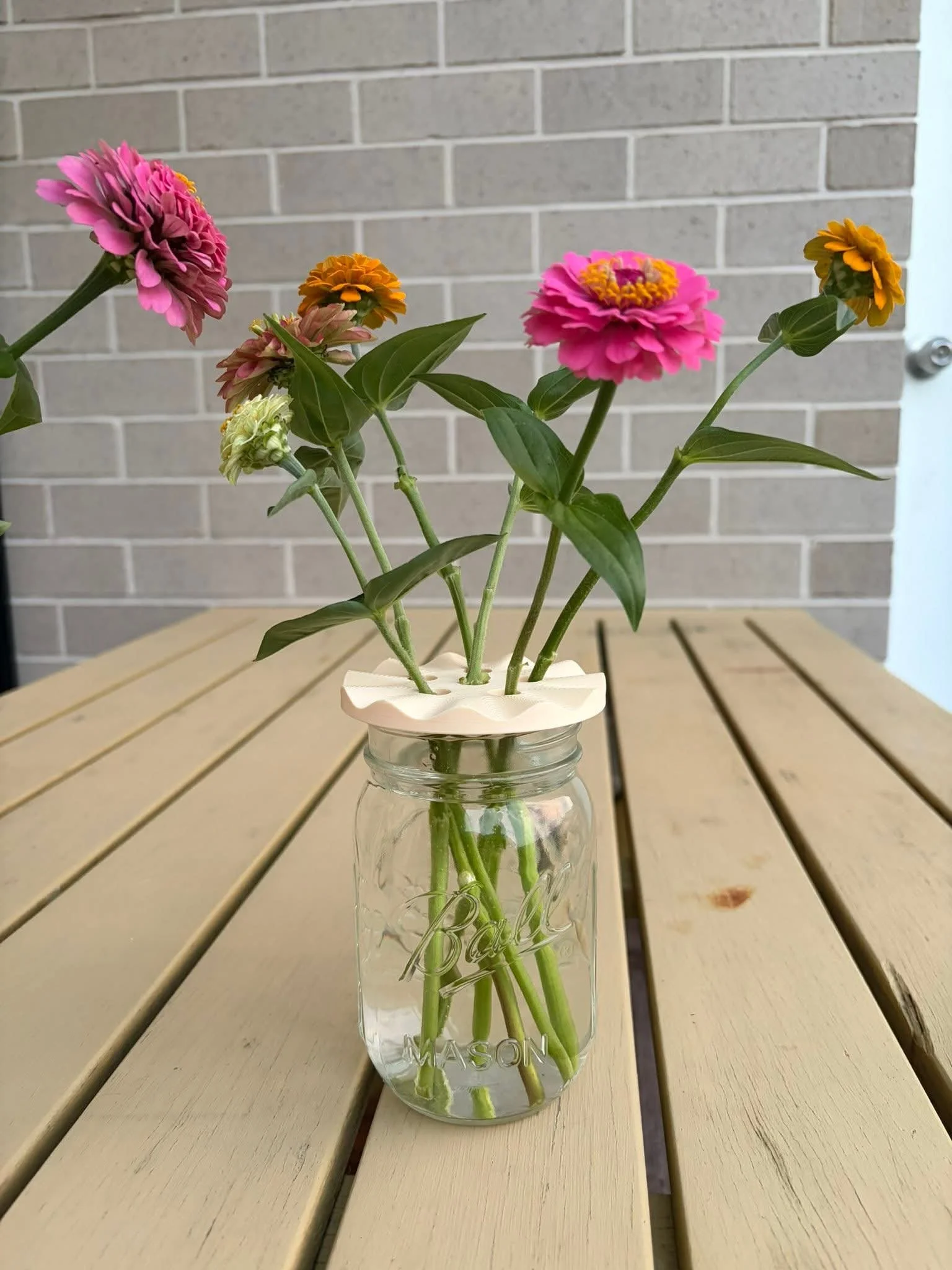 A glass mason jar with water and green flower stems, holding a bouquet of pink, orange, and white flowers with green leaves, placed on a wooden table with a brick wall in the background.