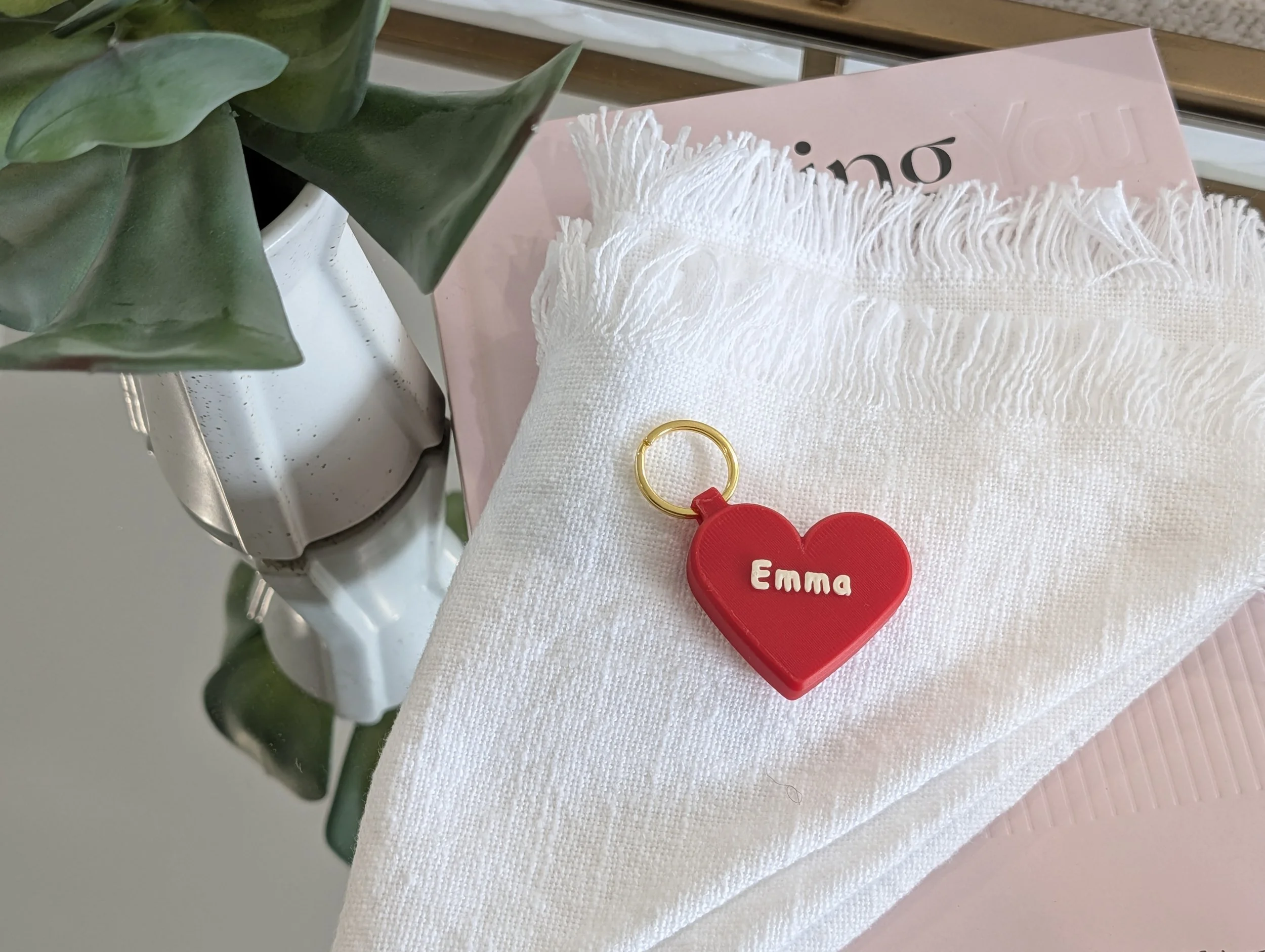 A white heart-shaped keychain with the name 'Emma' on it, resting on a white textured cloth, next to a green plant in a white pot, with a pink book underneath the cloth.