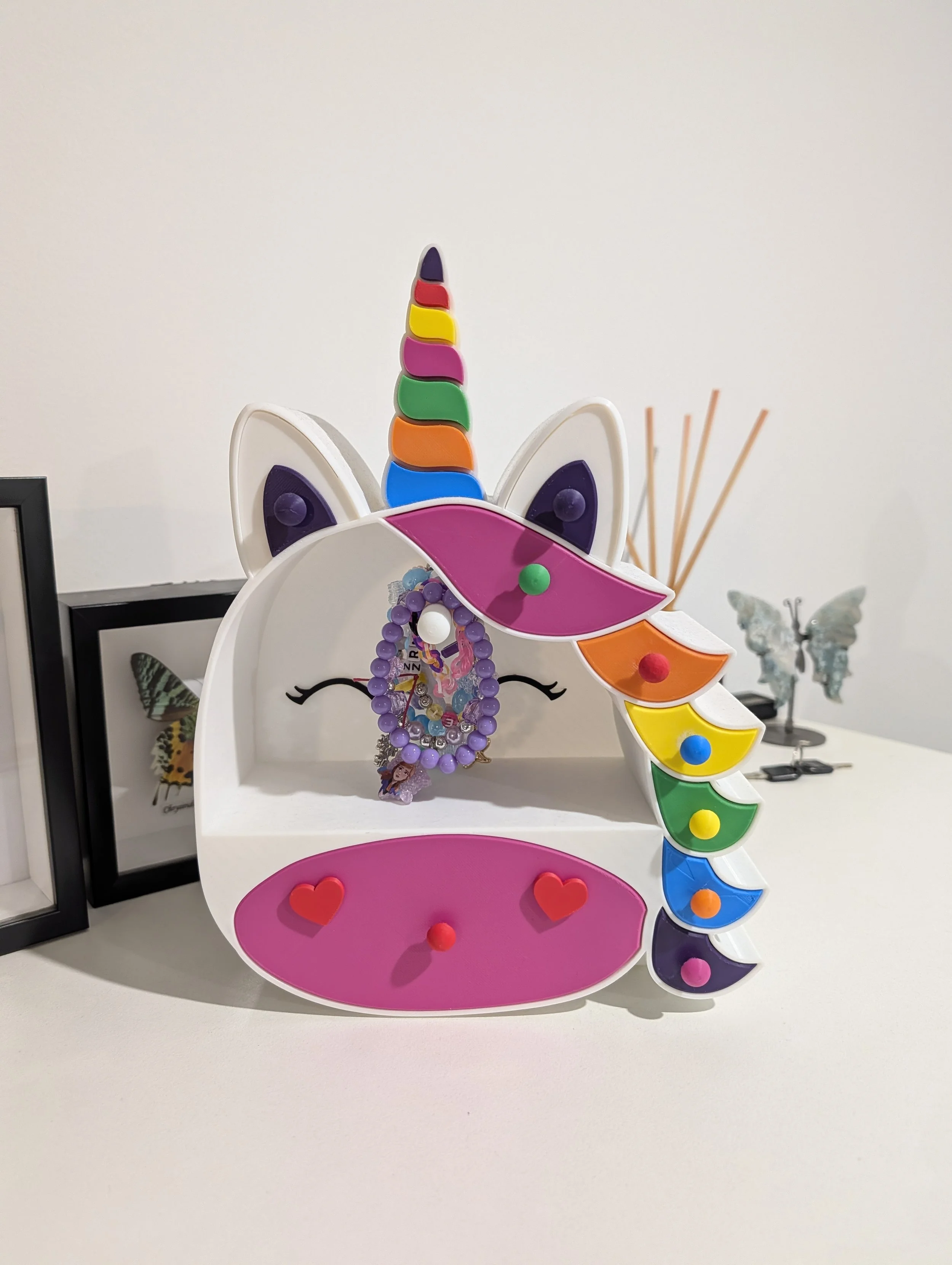 Unicorn-shaped jewelry holder with a rainbow-colored horn and rainbow mane, holding necklaces, on a white surface, with butterflies and framed pictures in the background.
