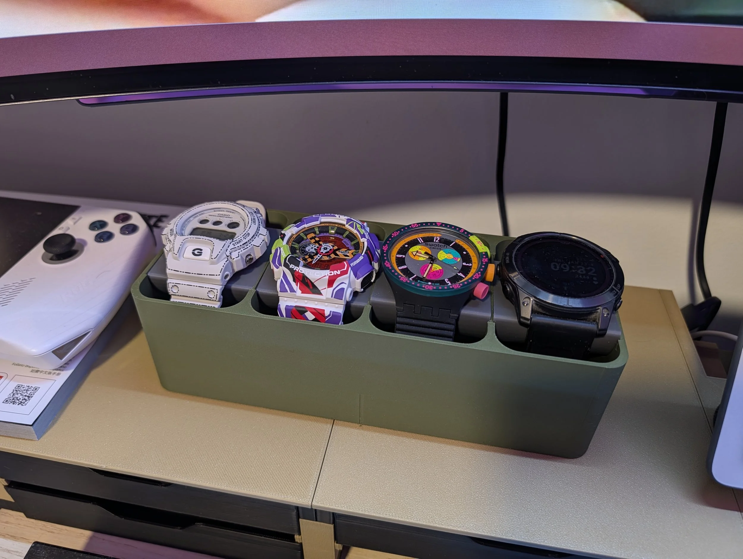 A collection of four watches in a green watch holder on a desk, with a game controller and a QR code on a book nearby, and a computer monitor above.