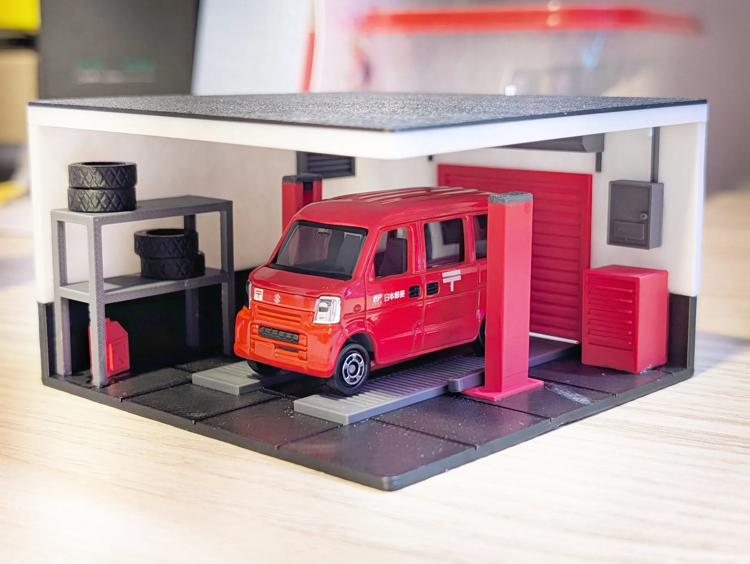 Miniature garage with a red Japanese postal delivery truck, shelves with tires, boxes, and garage equipment, all in a diorama setting.