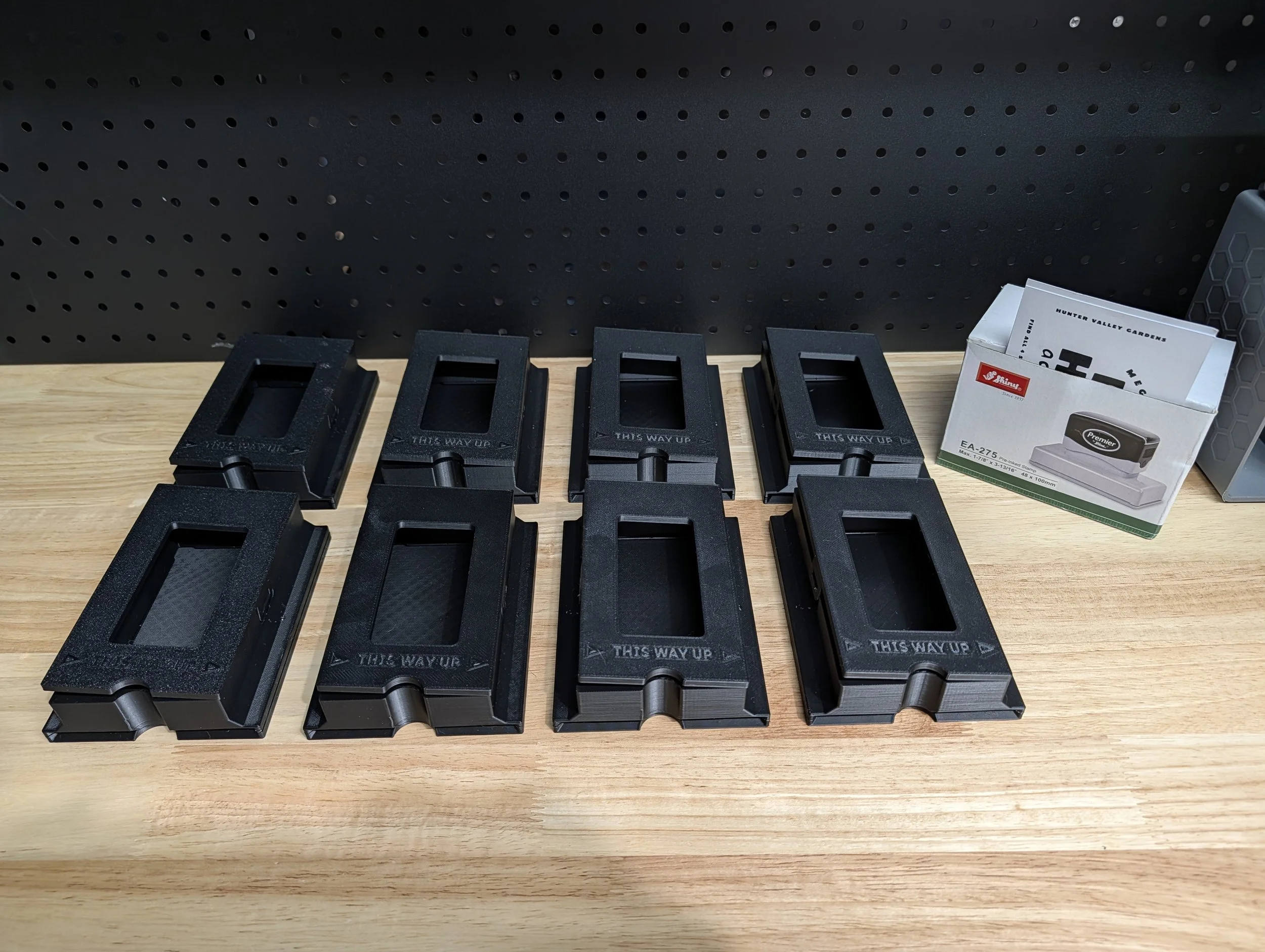 Eight black plastic injection mold holders on a wooden workbench, with a box of paper punches labeled 'Hunter Valley Gardens' and 'Premier' to the right.