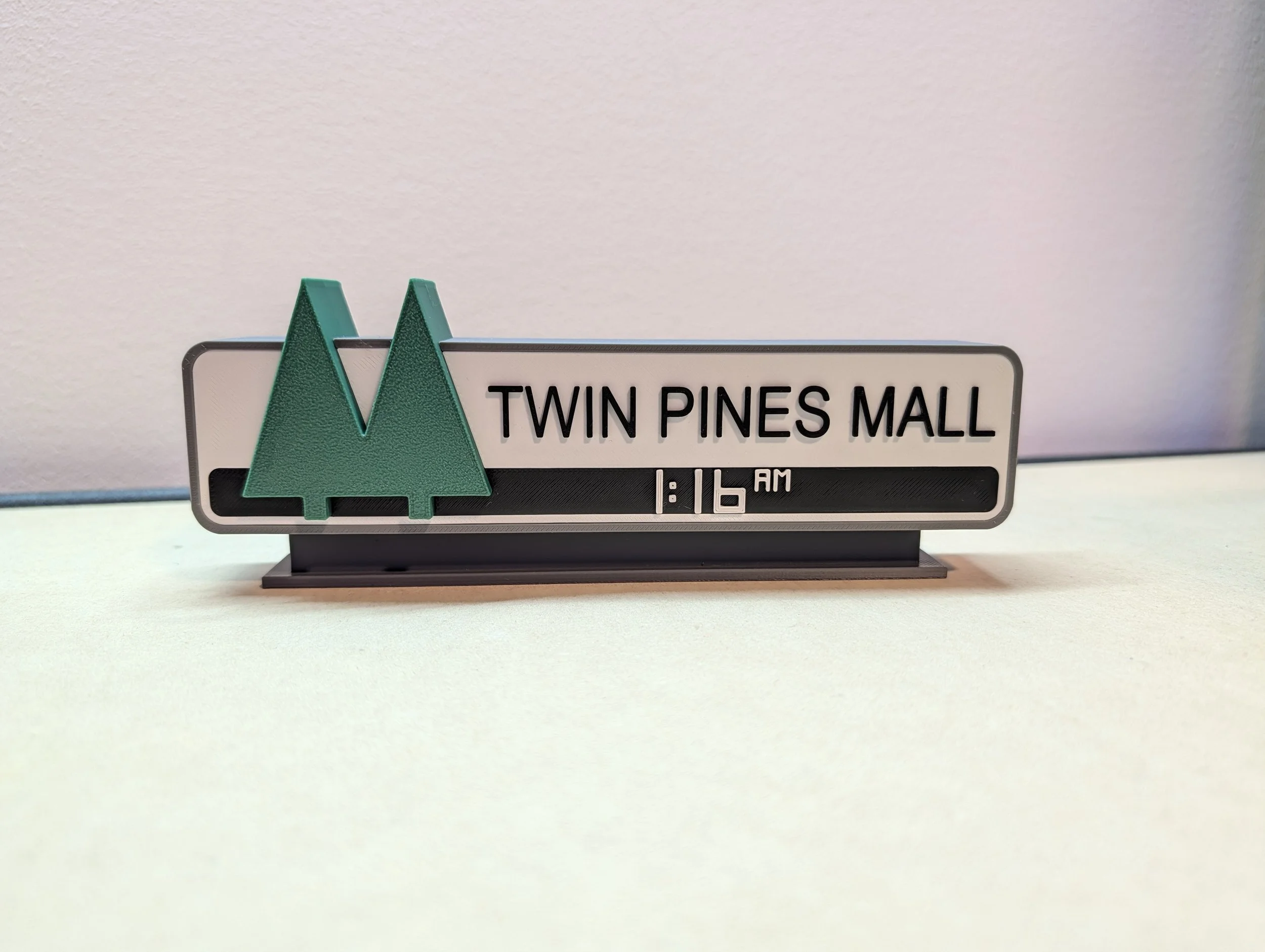 A digital desk sign displaying "Twin Pines Mall" with a clock showing 1:16 AM. The sign has two green pine tree icons on the left side and is placed on a light-colored surface against a light wall.