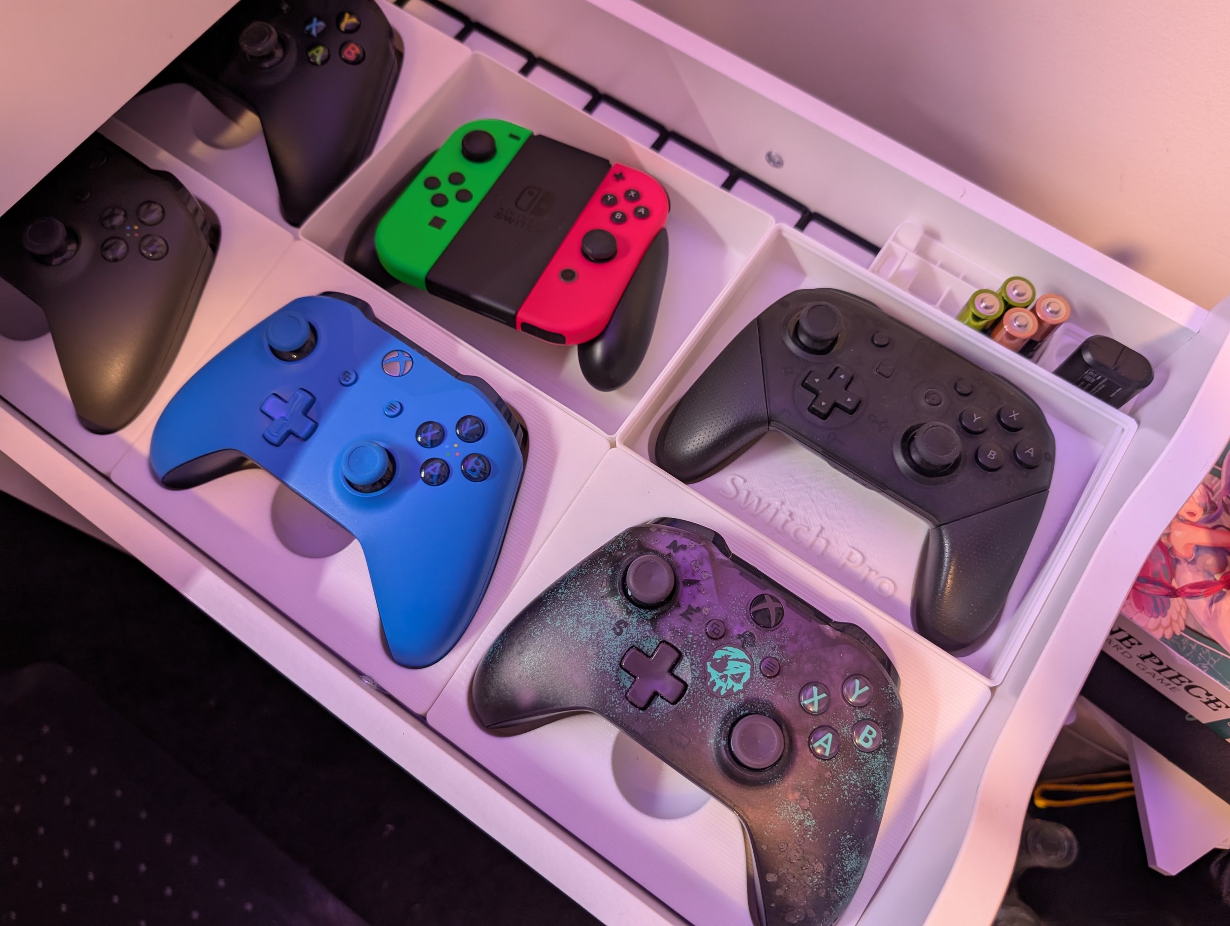 A collection of various gaming controllers, including Xbox and Nintendo Switch controllers, arranged in a white box with compartments, along with batteries and a charger.