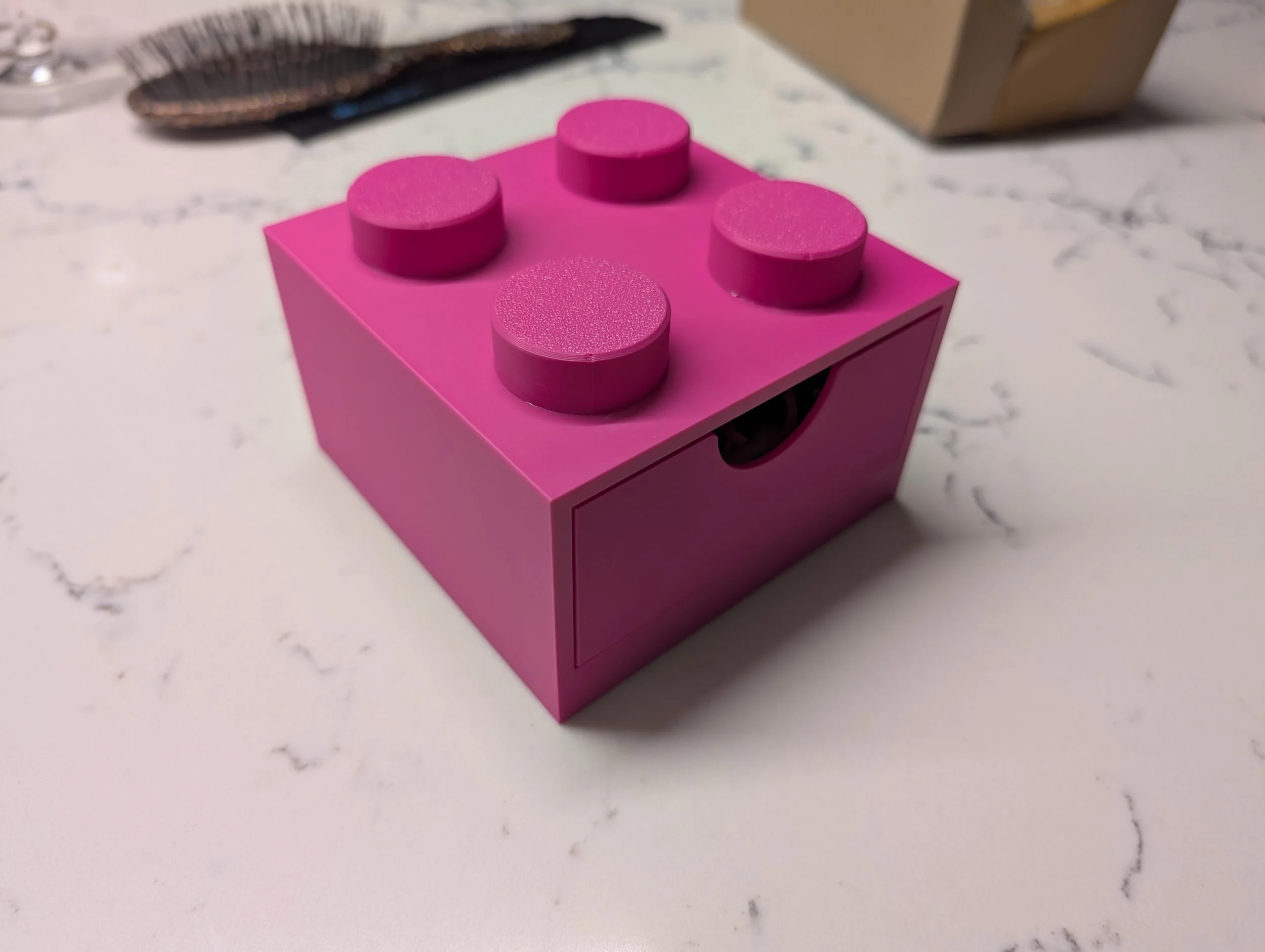 Pink LEGO-style box with four round studs on top, placed on a white marble surface.