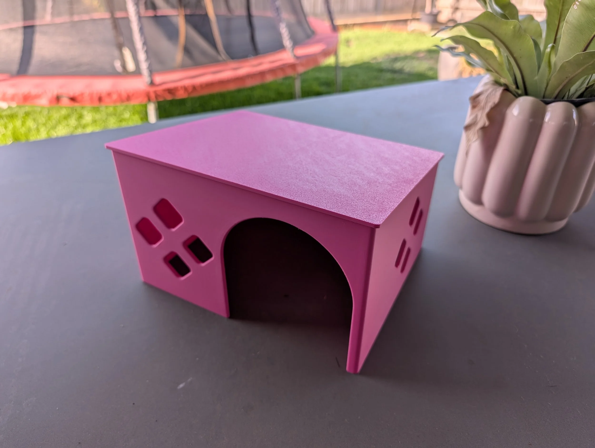 Small pink wooden house-shaped object with cutouts on the sides, placed on a gray surface outdoors. There is a white pot with green plants to the right and a red trampoline with a safety net in the background.