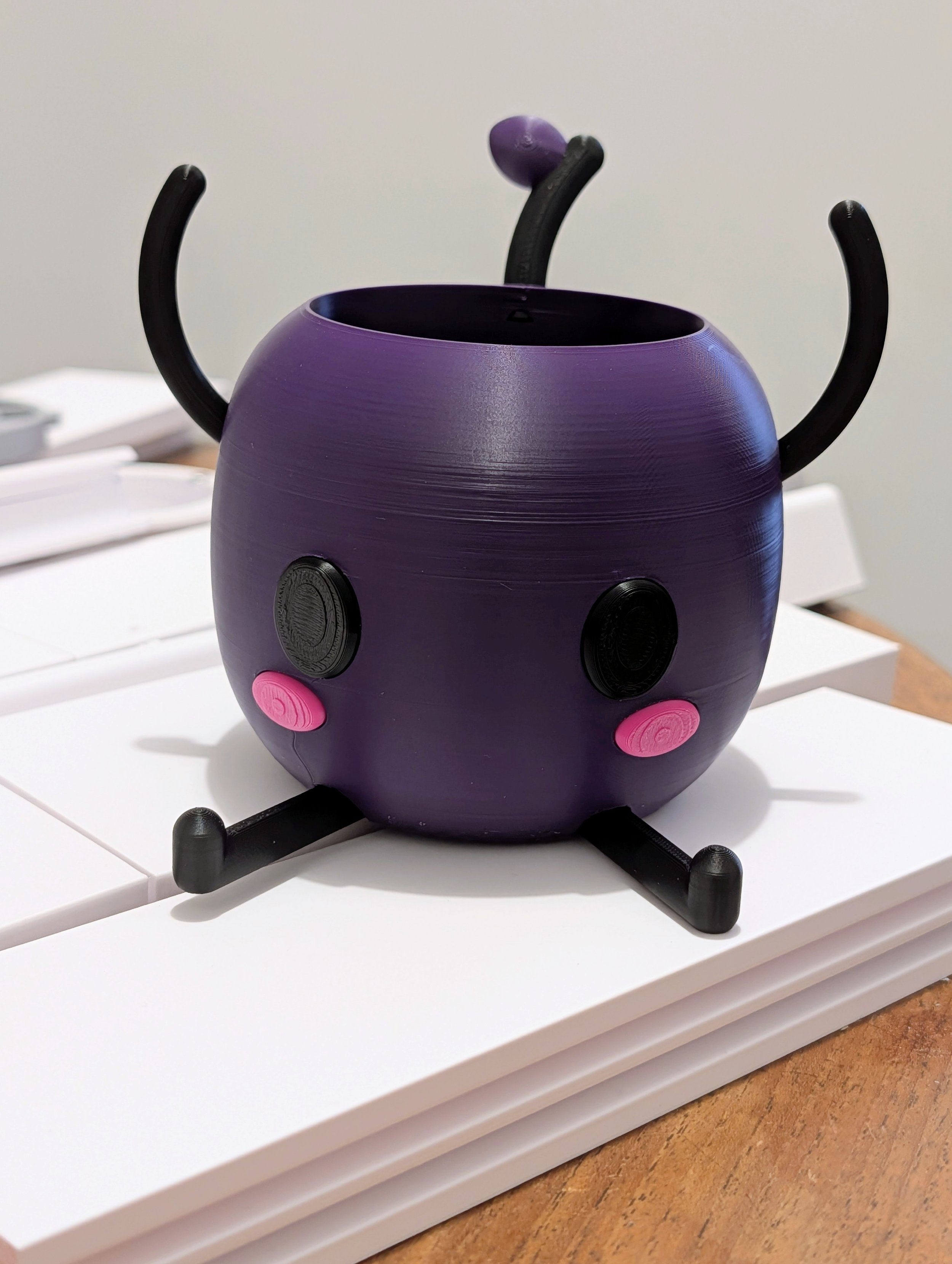 A creative cat-shaped object with a purple body, black legs, and pink cheeks, placed on a white surface.
