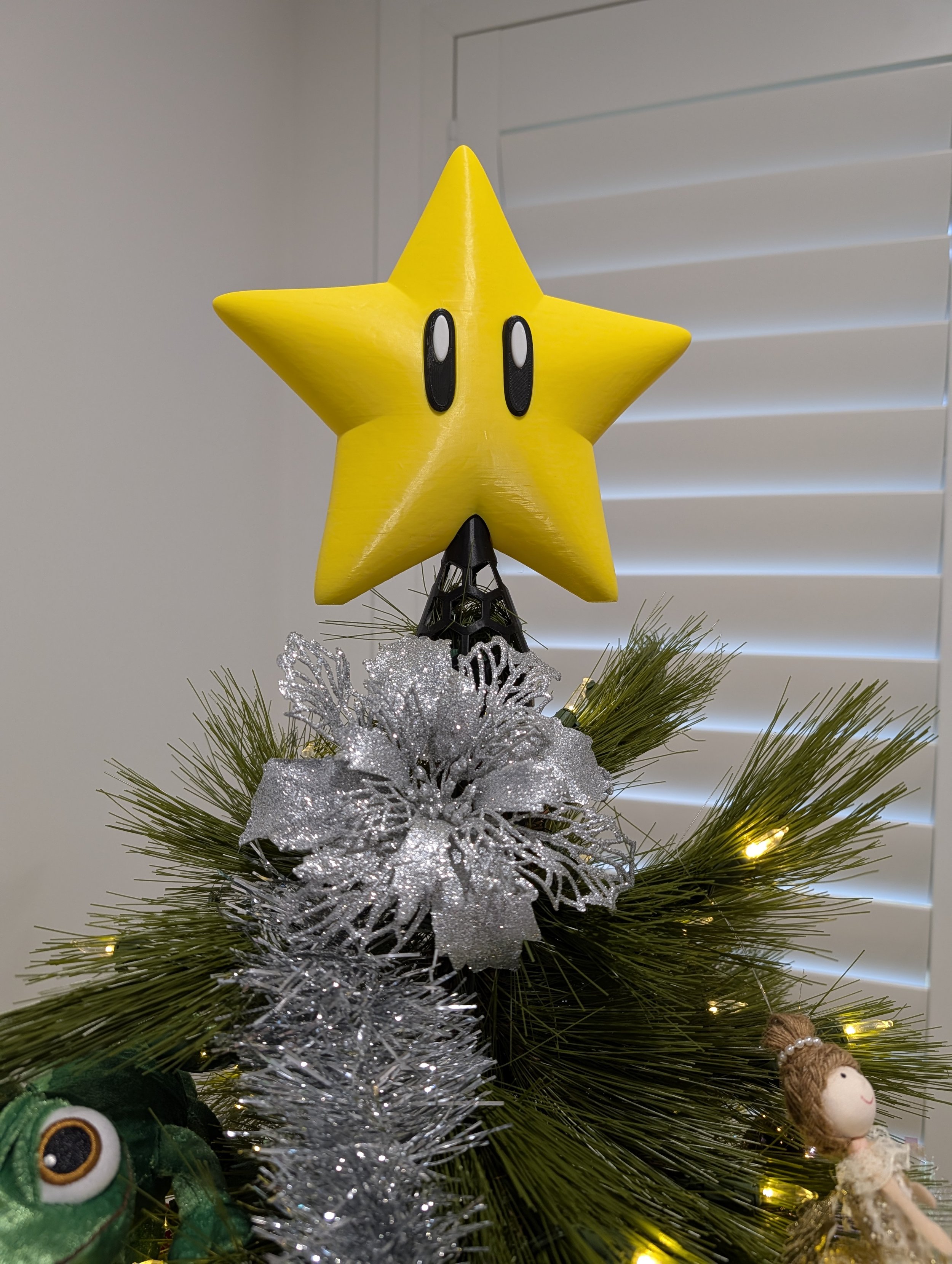 Decorated Christmas tree with a yellow star topper featuring black eyes and a black base, silver ornaments, greenery, small lights, and a doll ornament with a ponytail.