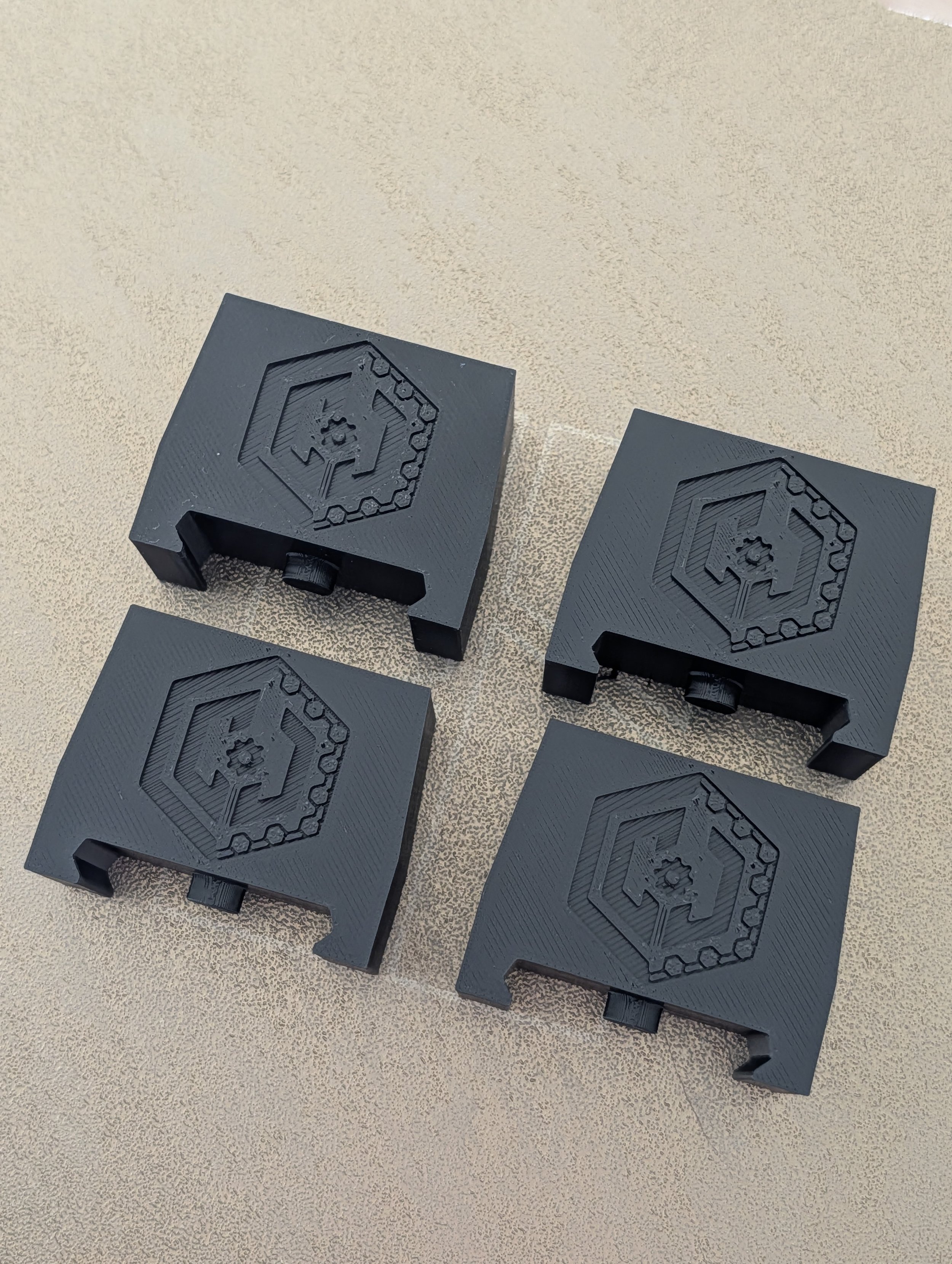 Four black plastic cable clips with a gear and chain icon embossed on top, placed on a beige textured surface.