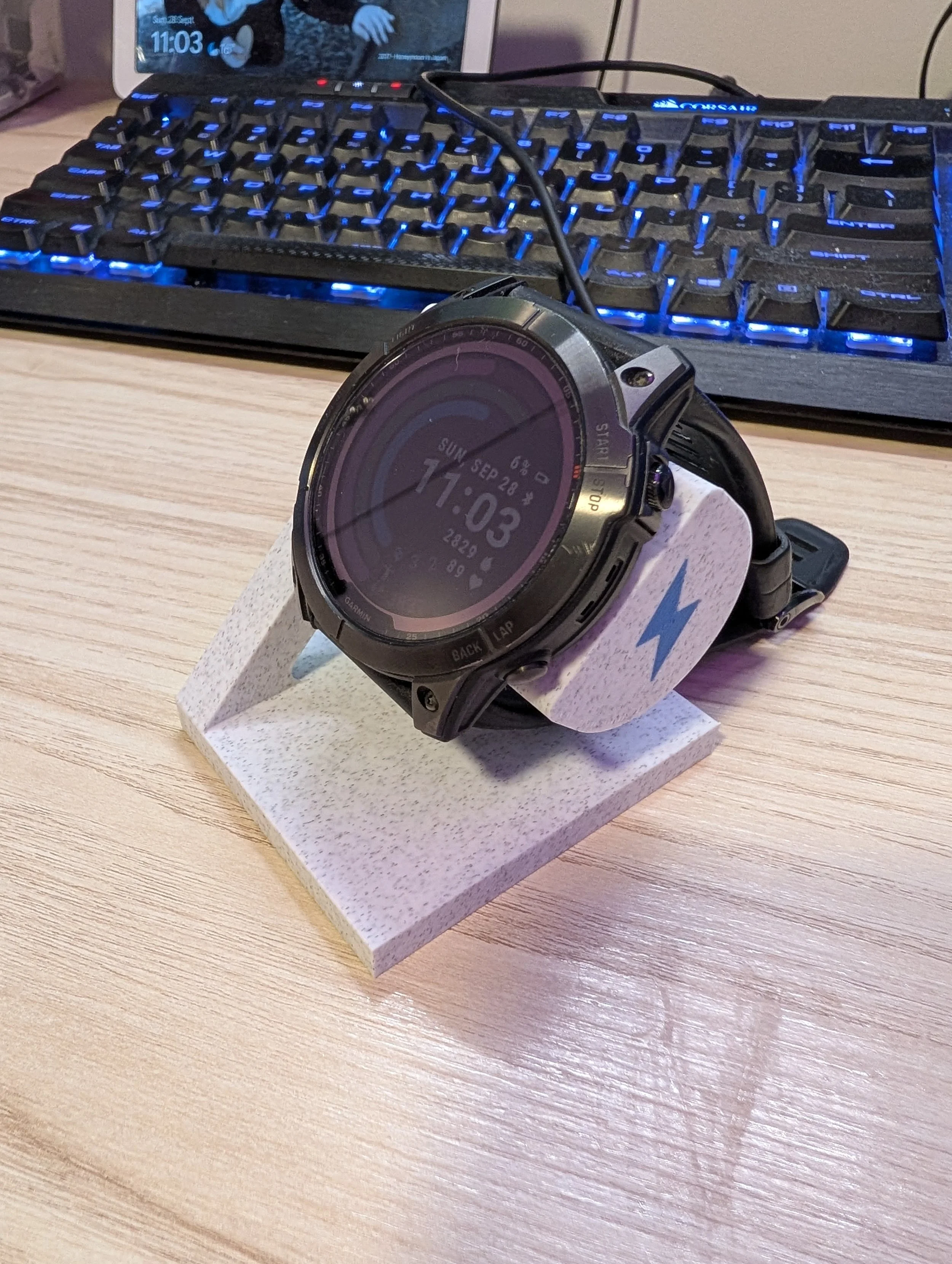 A smartwatch placed on a white stand with a blue lightning bolt symbol, on a wooden desk. In the background, there is a black keyboard with blue backlit keys and a computer monitor showing the time 11:03.