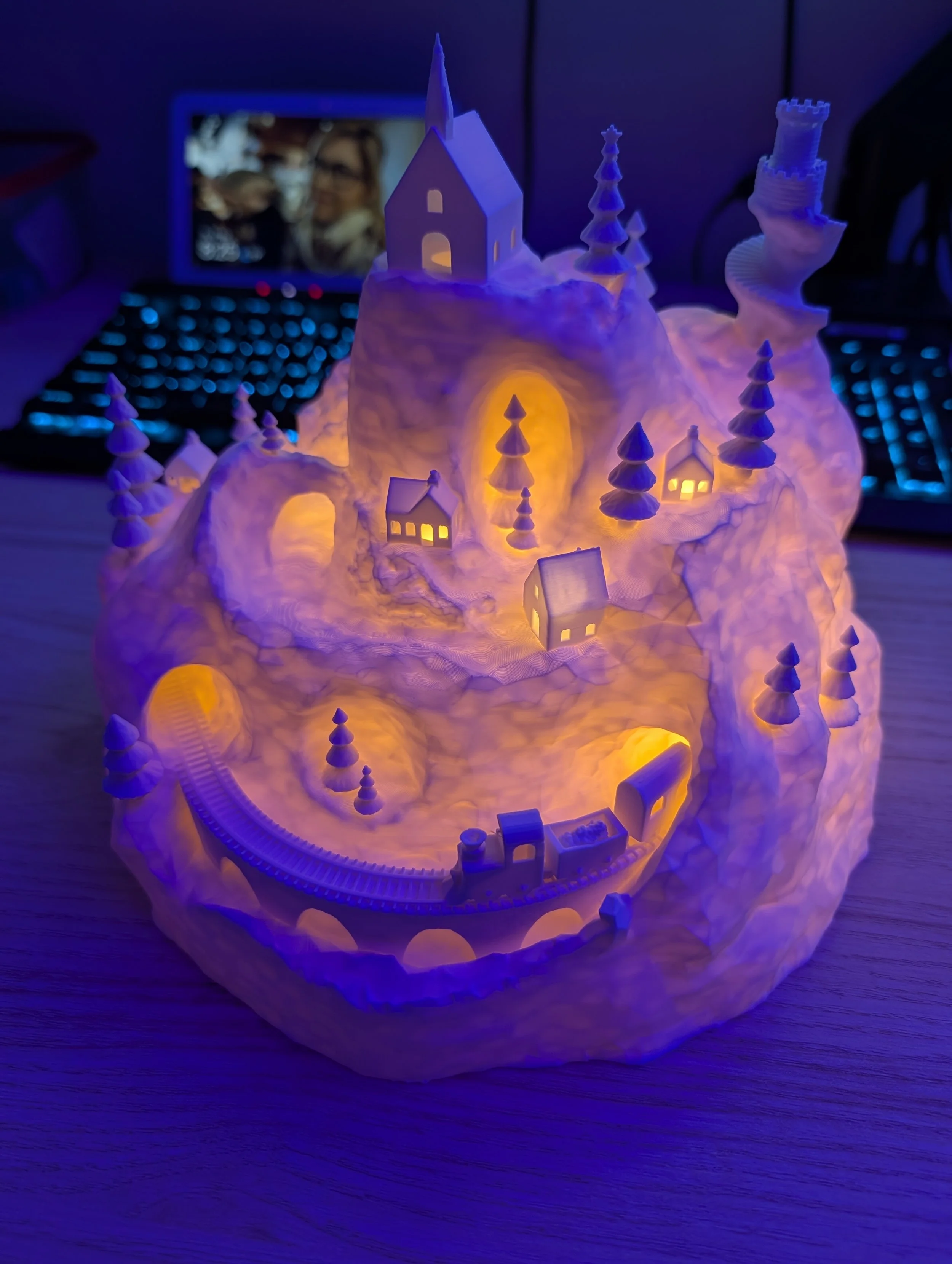 Illuminated snow mountain scene with miniature houses, trees, a train, and a church, glowing in blue and yellow lights.