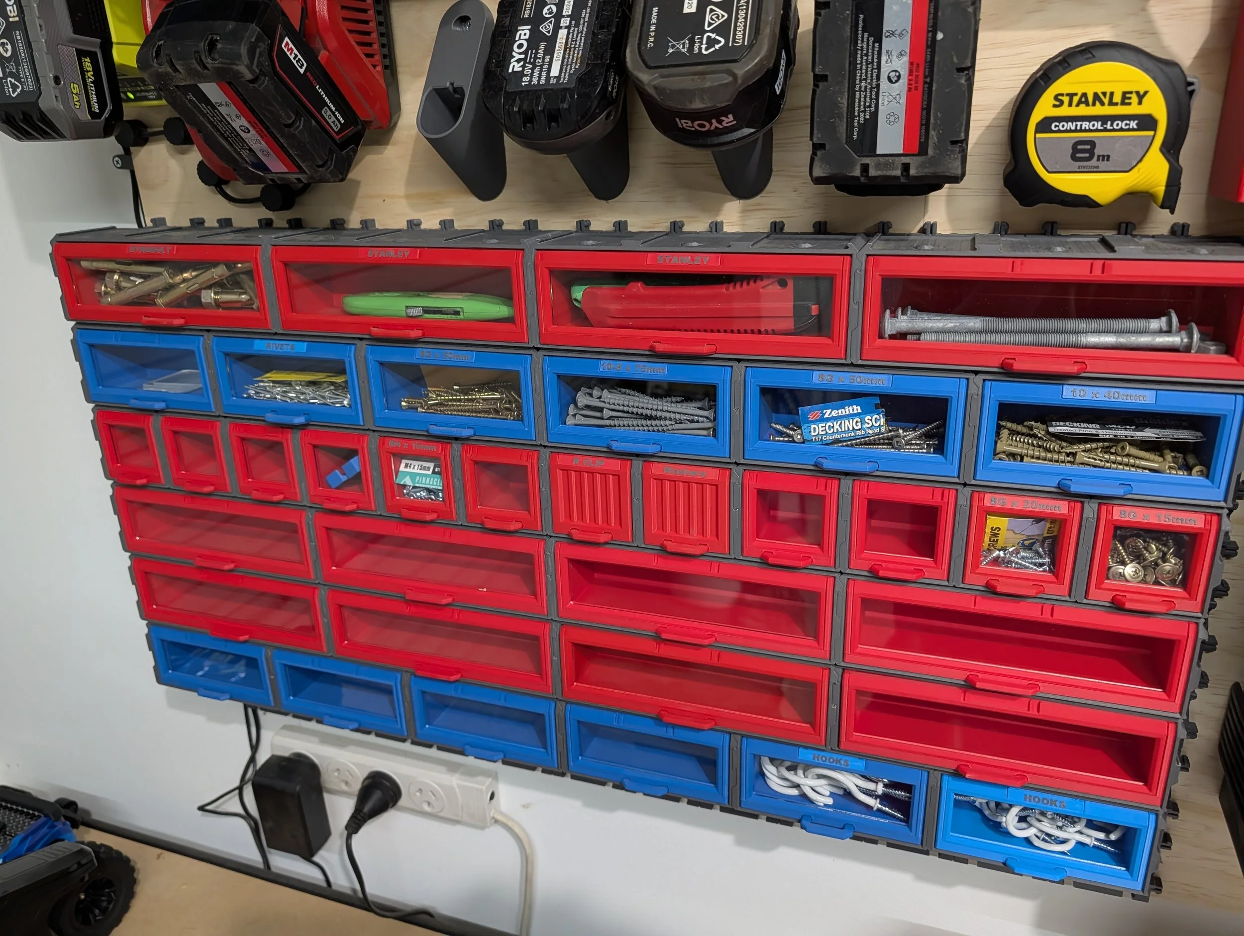 Organized workshop storage with red and blue plastic drawers containing screws, nails, hooks, and hardware, mounted on a wall below power tools and a measuring tape.
