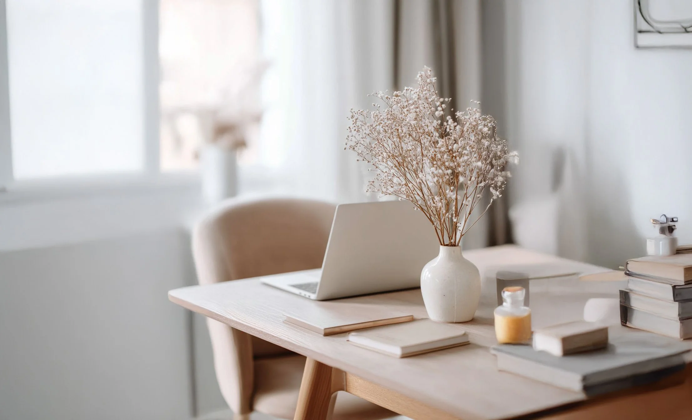 Un bureau en bois clair avec un vase blanc contenant des fleurs séchées, un ordinateur portable, plusieurs livres empilés, un cahier et des flacons de crème ou de lotion, dans une pièce lumineuse.