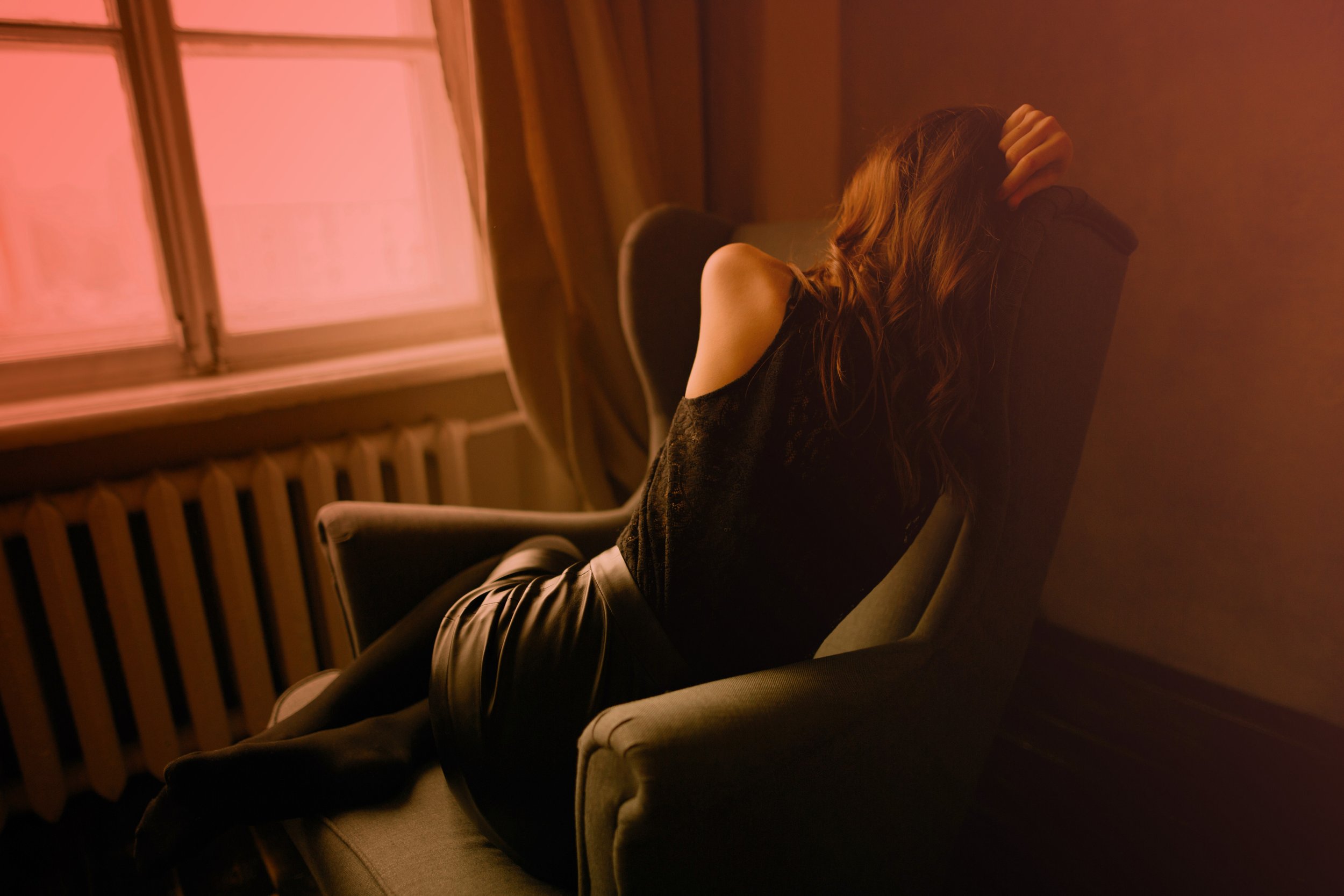 Photographie d'une femme vue de dos, assise dans un fauteuil confortable près d'une fenêtre. L'ambiance tamisée et sa posture suggèrent la fatigue, l'introspection et le besoin d'apaisement émotionnel face au stress quotidien.